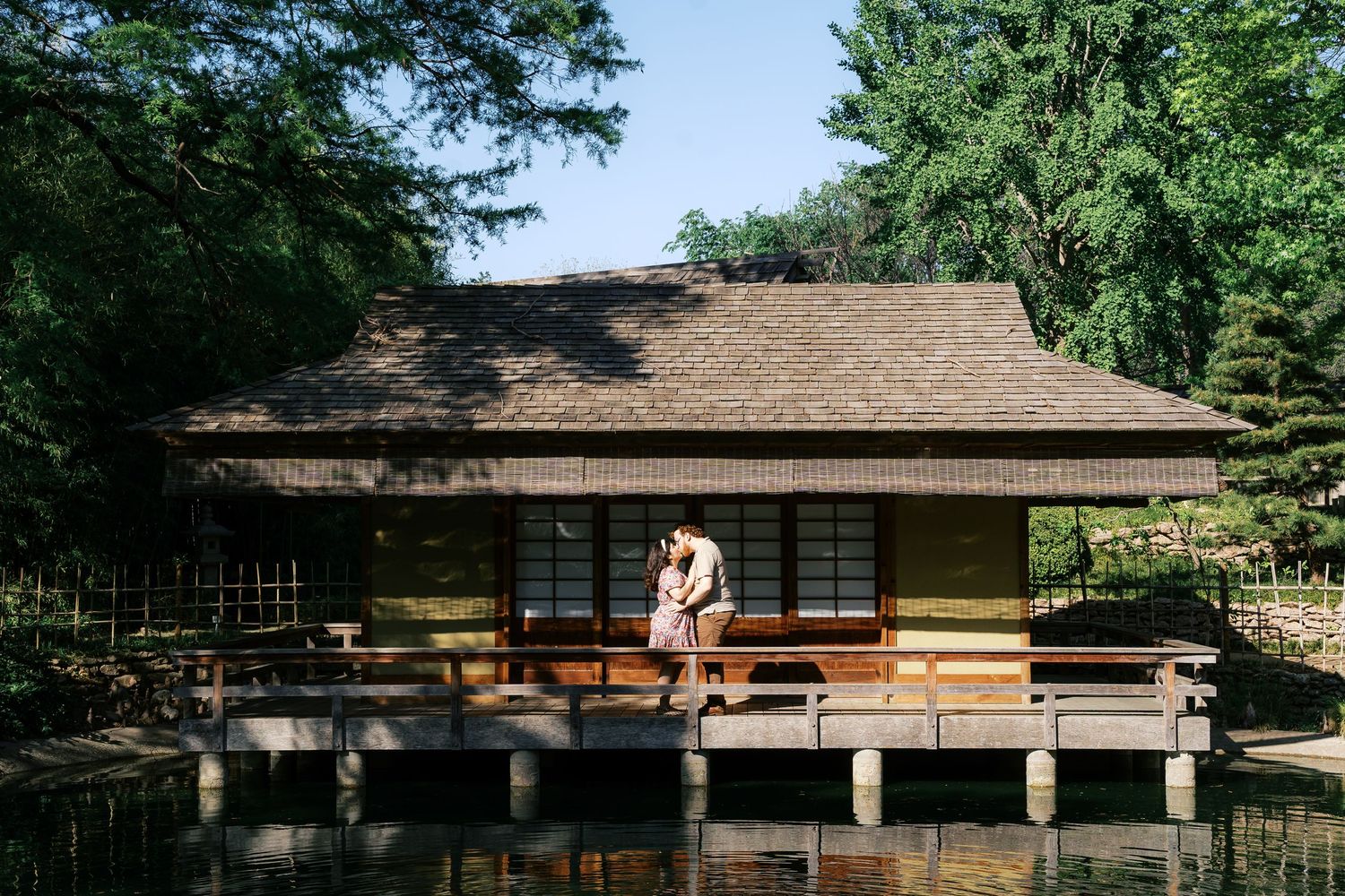 Couple embracing on wooden deck of traditional Japanese garden teahouse surrounded by lush trees and serene pond.