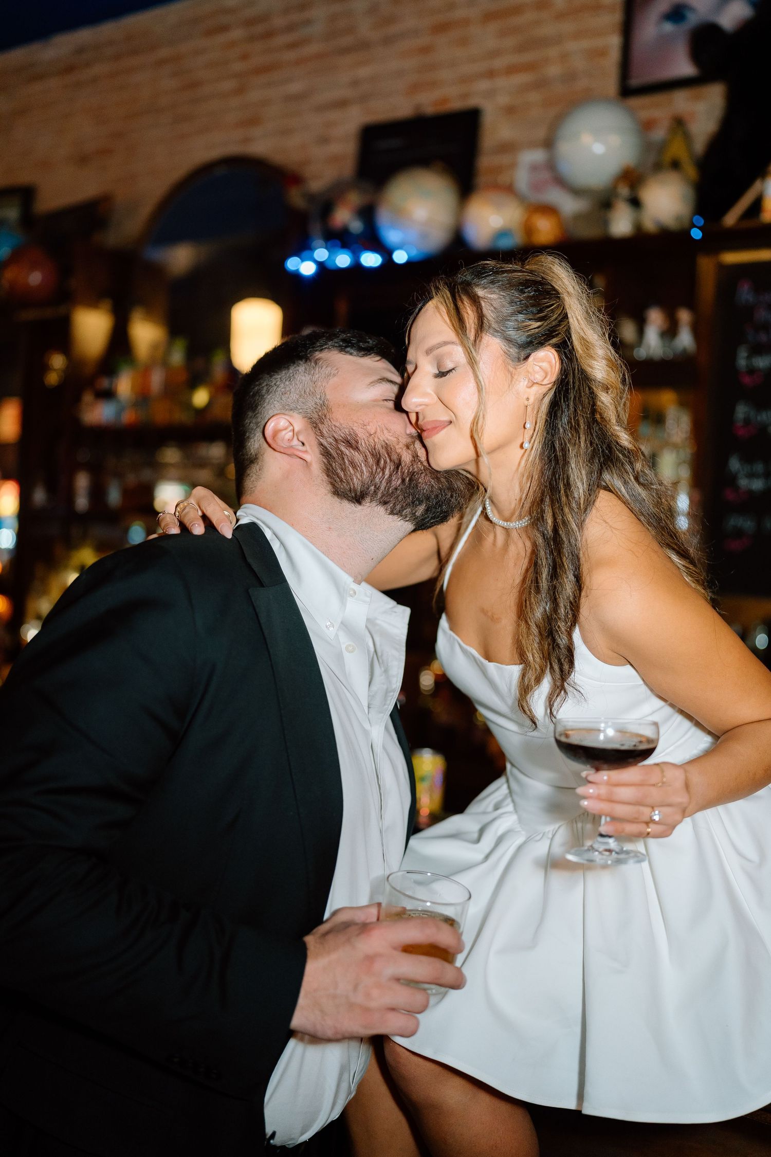 Couple in formal wear shares tender moment while holding wine glass at bar.
