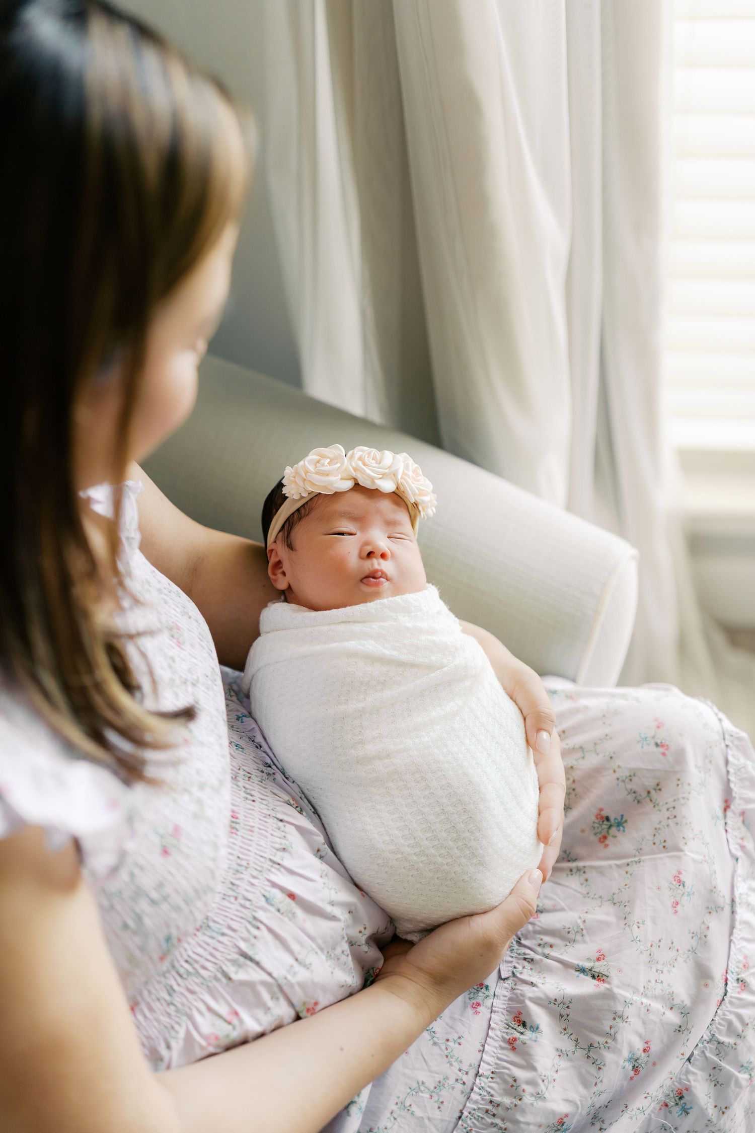 A newborn baby wrapped in white swaddle blanket rests peacefully against floral fabric near a window.