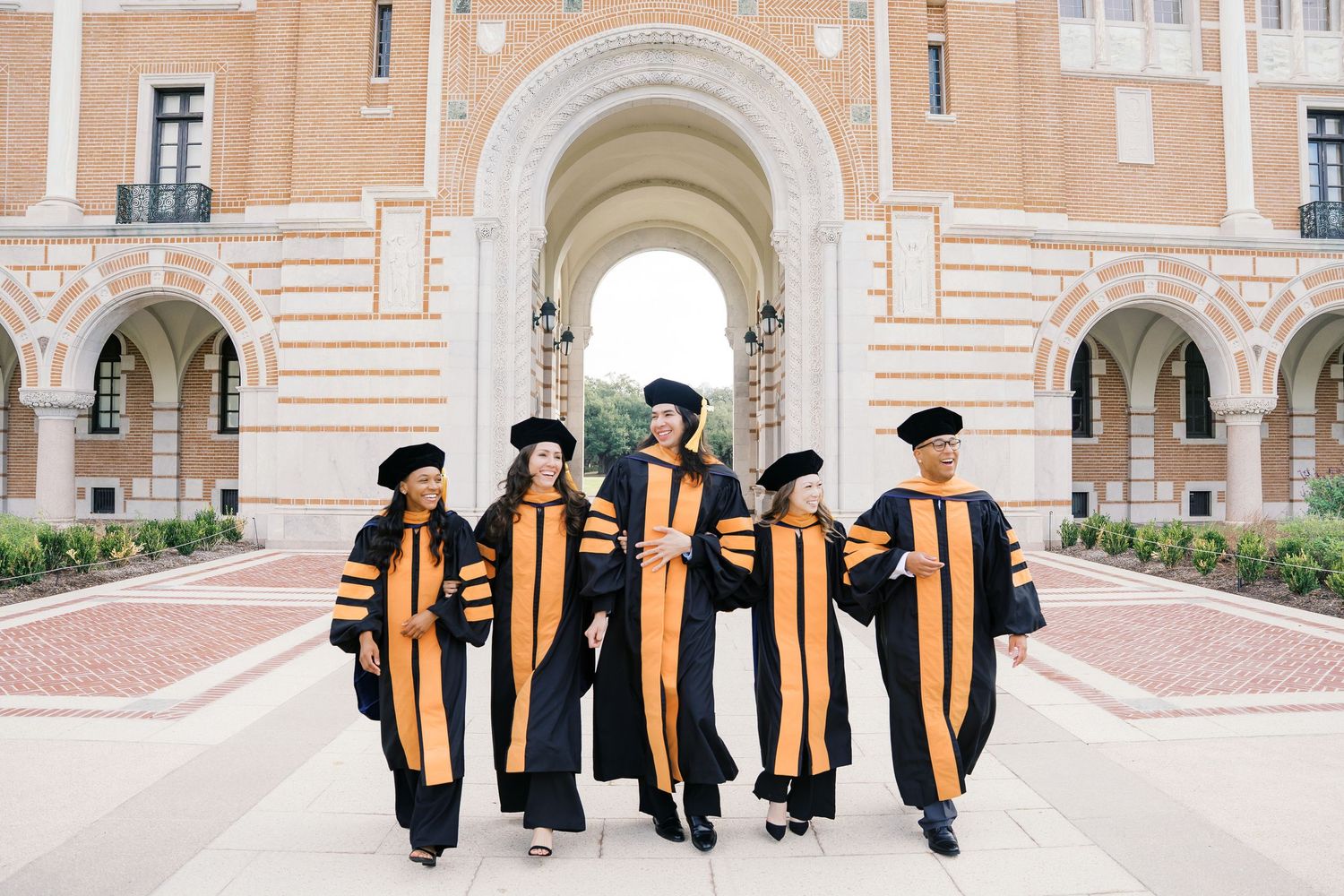 A group of graduates in black and gold academic regalia walking along a brick building during commencement ceremony.