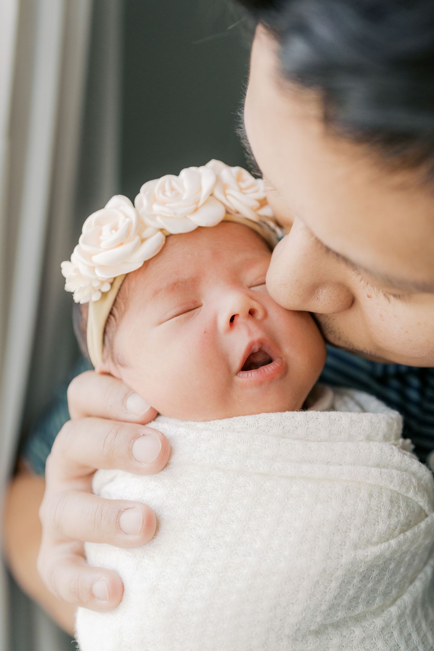 Intimate close-up of parent giving newborn baby wearing floral headband a gentle kiss.