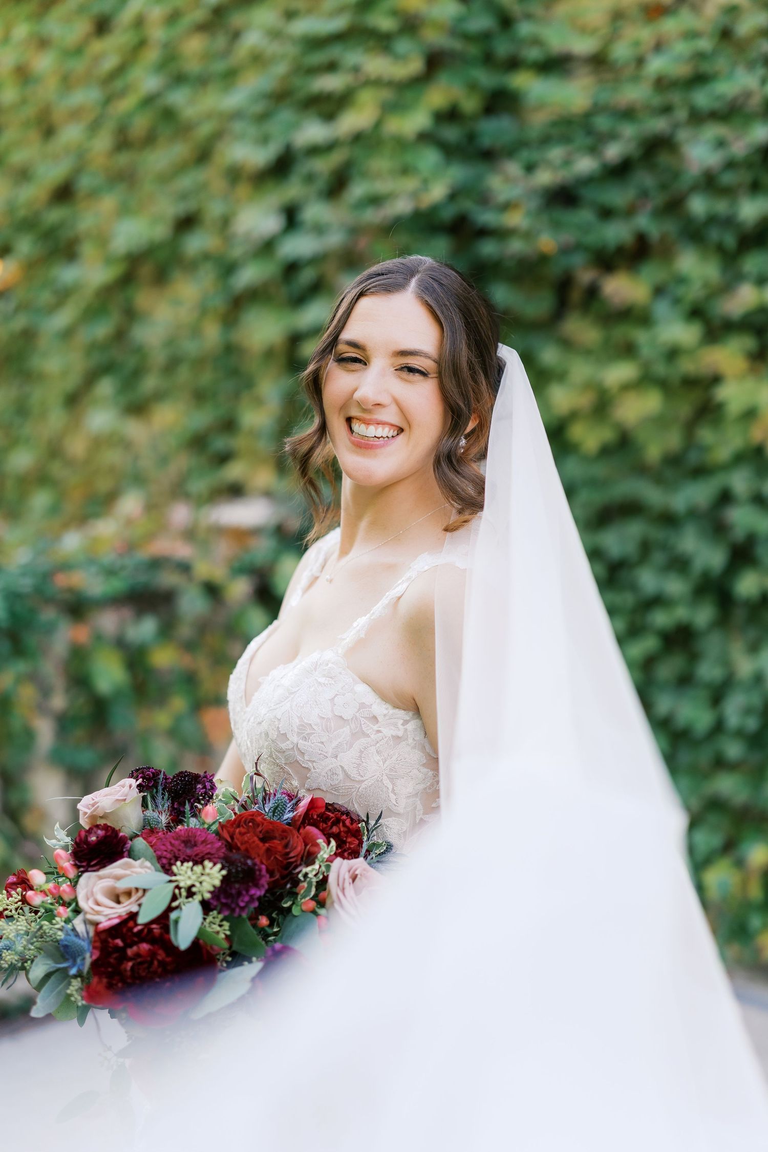 Bride in white lace gown smiles while holding deep burgundy and blush rose bouquet.