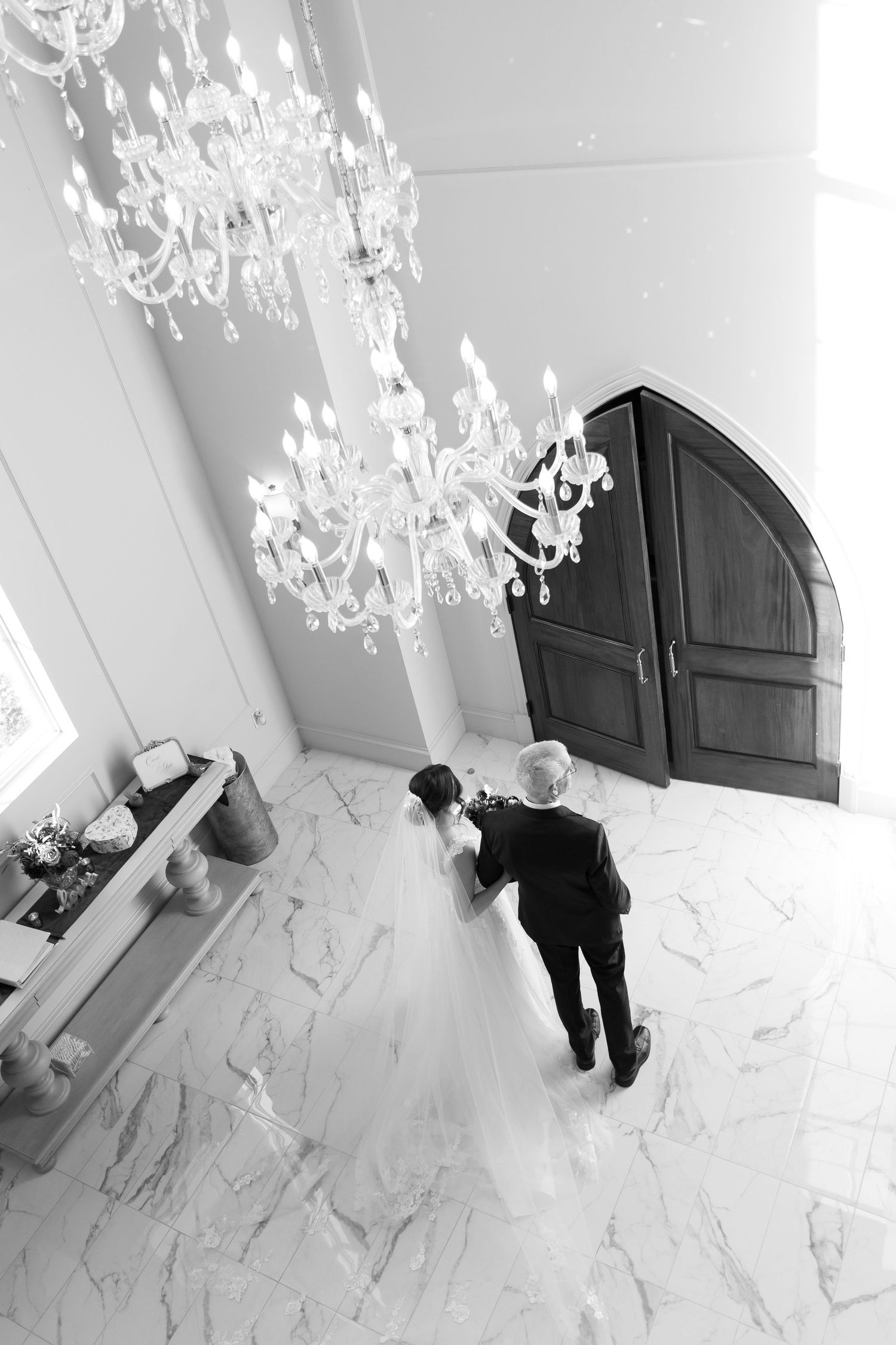 Black and white image of elegant crystal chandeliers hanging in a marble-floored hallway.
