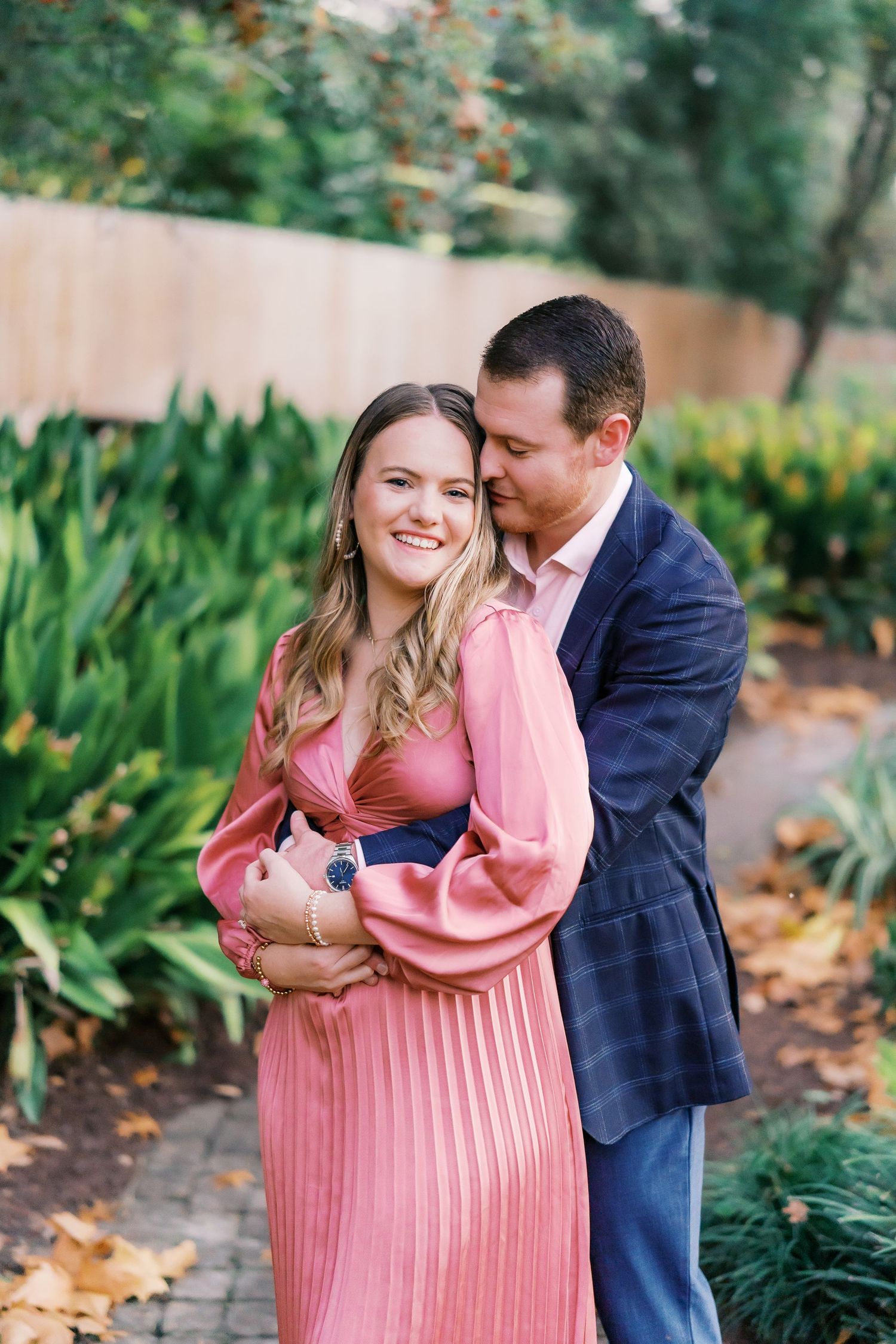 A couple poses for engagement photos in a lush garden setting wearing navy and pink.