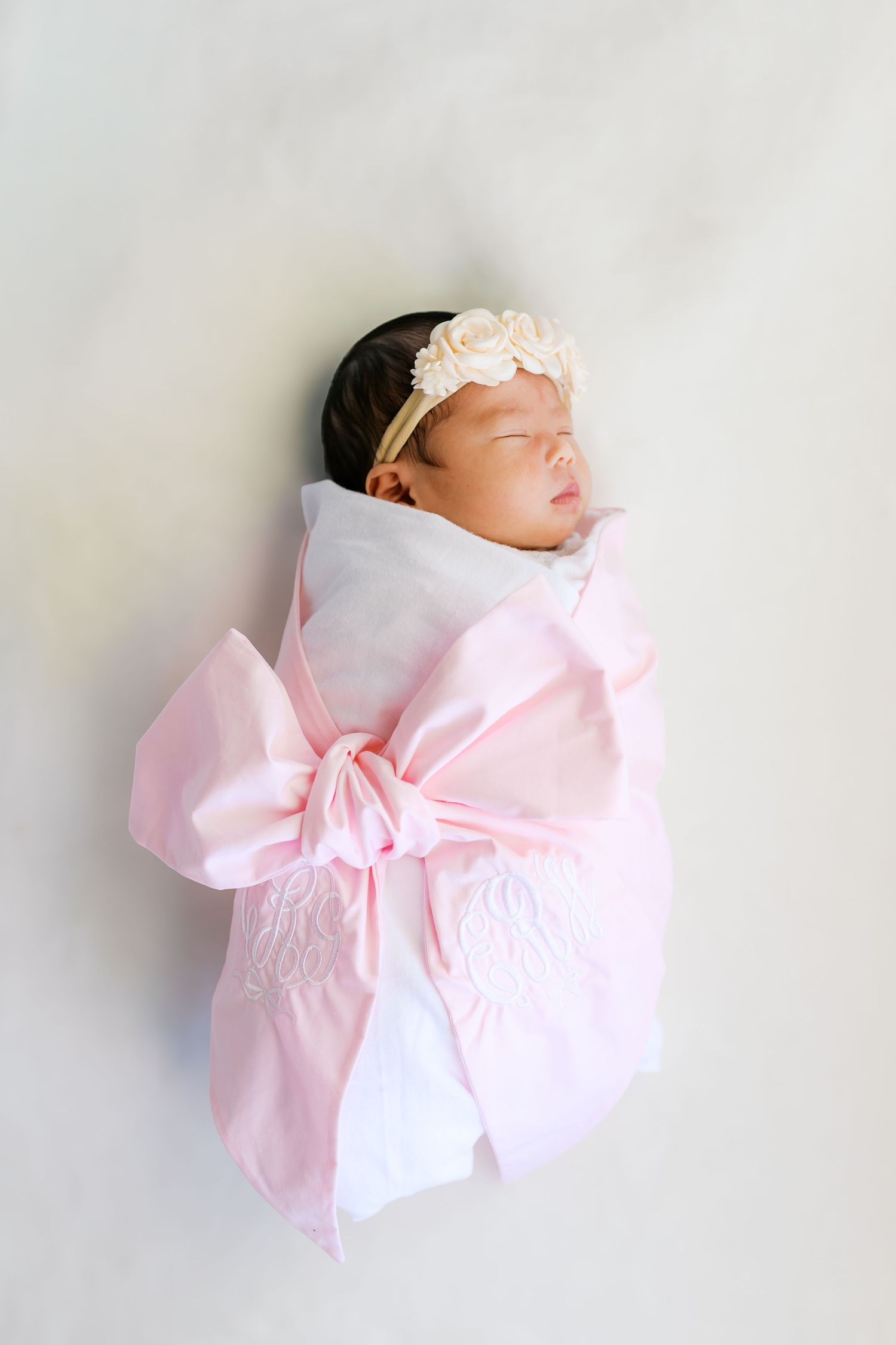 Sleeping newborn wrapped in white and pink bow swaddle with floral headband against white background.