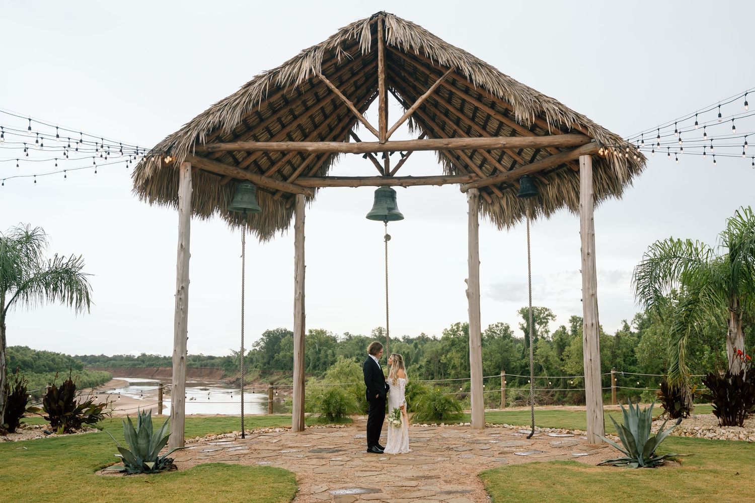 A thatched gazebo with string lights and a bell overlooking a scenic waterfront setting.