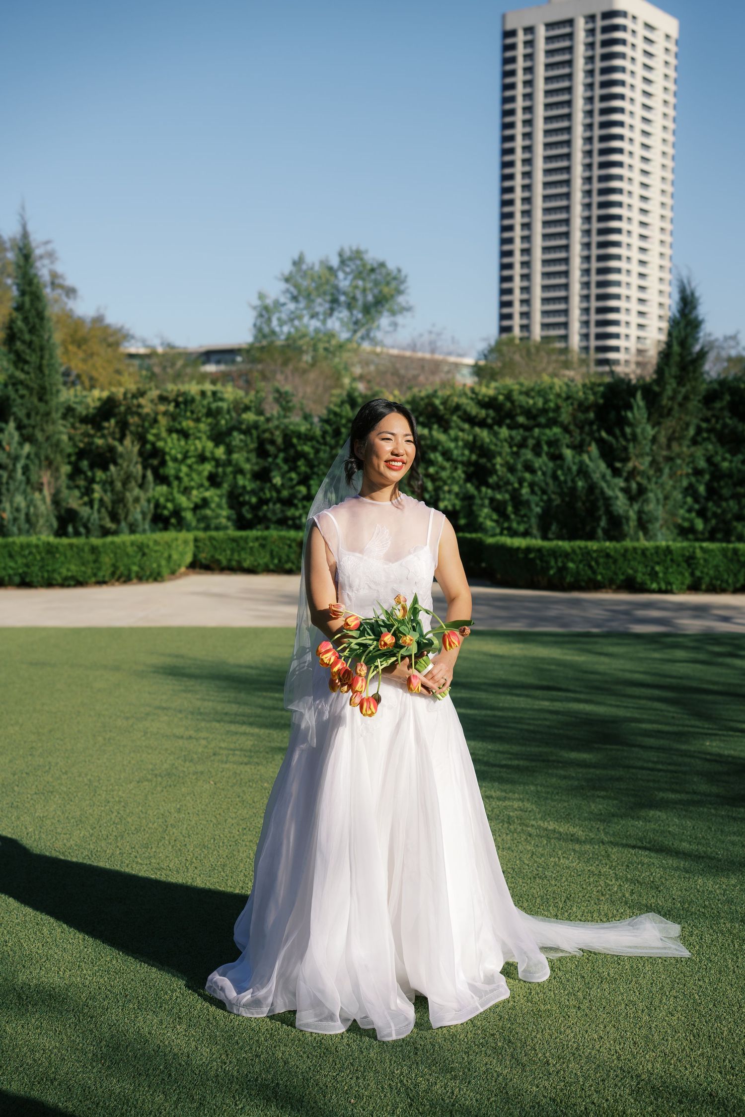 Smiling bride in white gown and veil holds colorful bouquet outdoors on green lawn with hedges and skyscraper behind.
