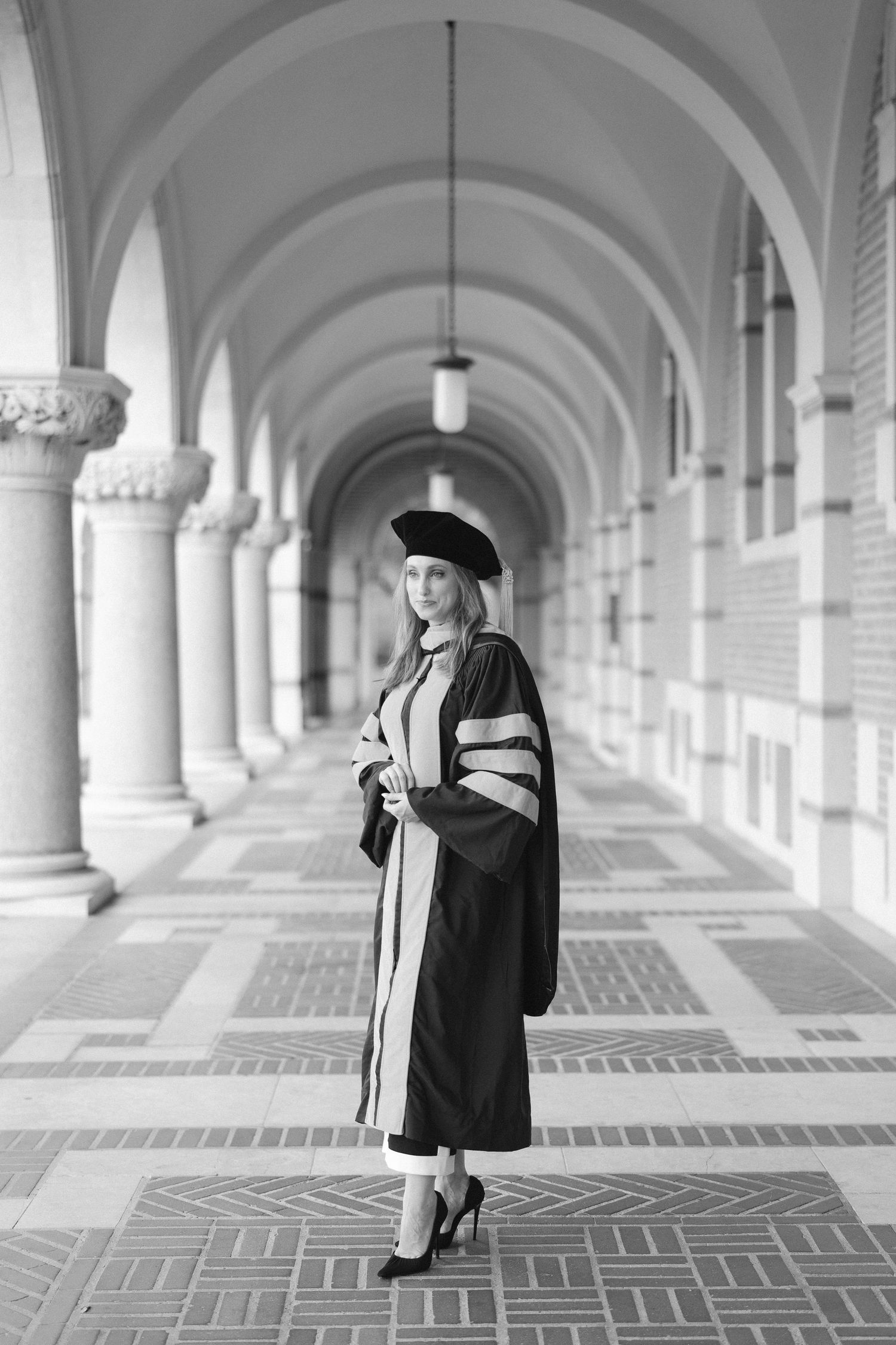 Black and white portrait of graduate in academic regalia standing in columned hallway.