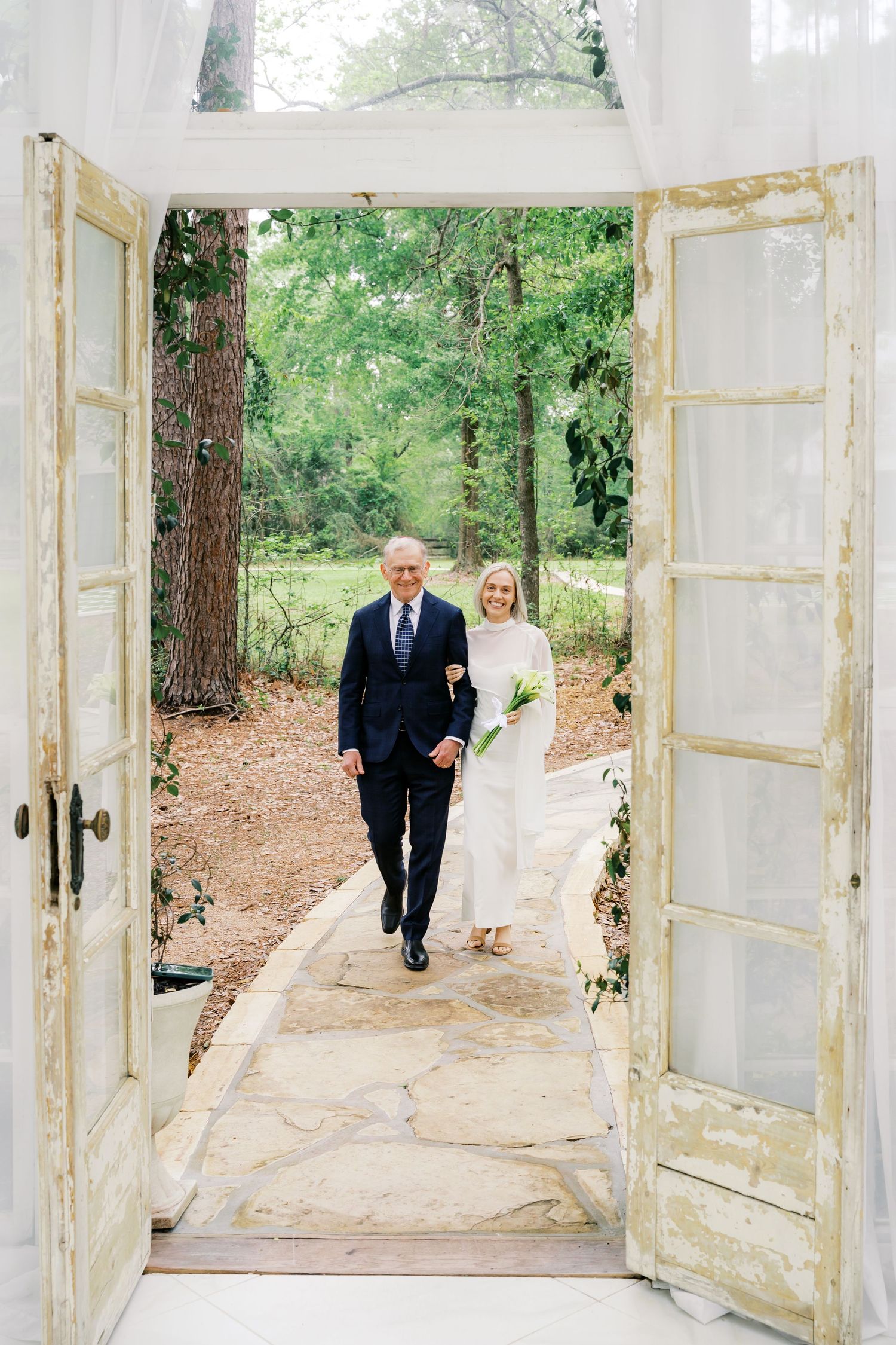 Bride in white gown walks with older man in navy suit through rustic French doors toward lush green woodland setting.