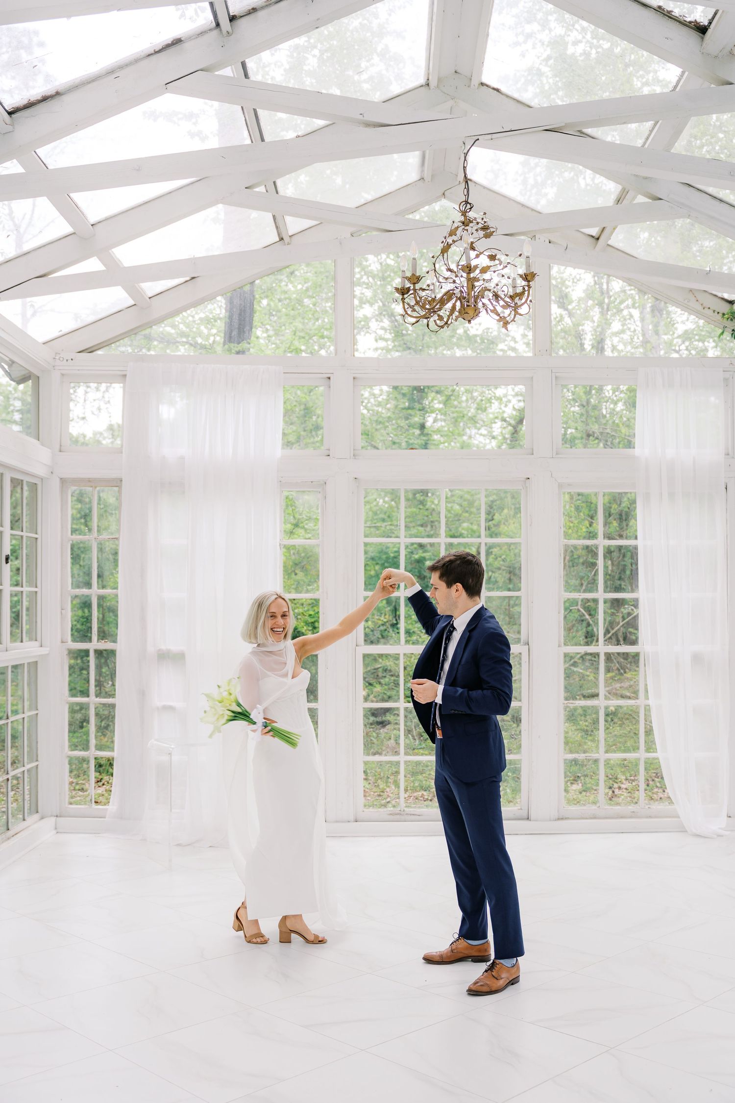 Bride and groom share first dance in elegant white greenhouse venue with chandelier and large windows.