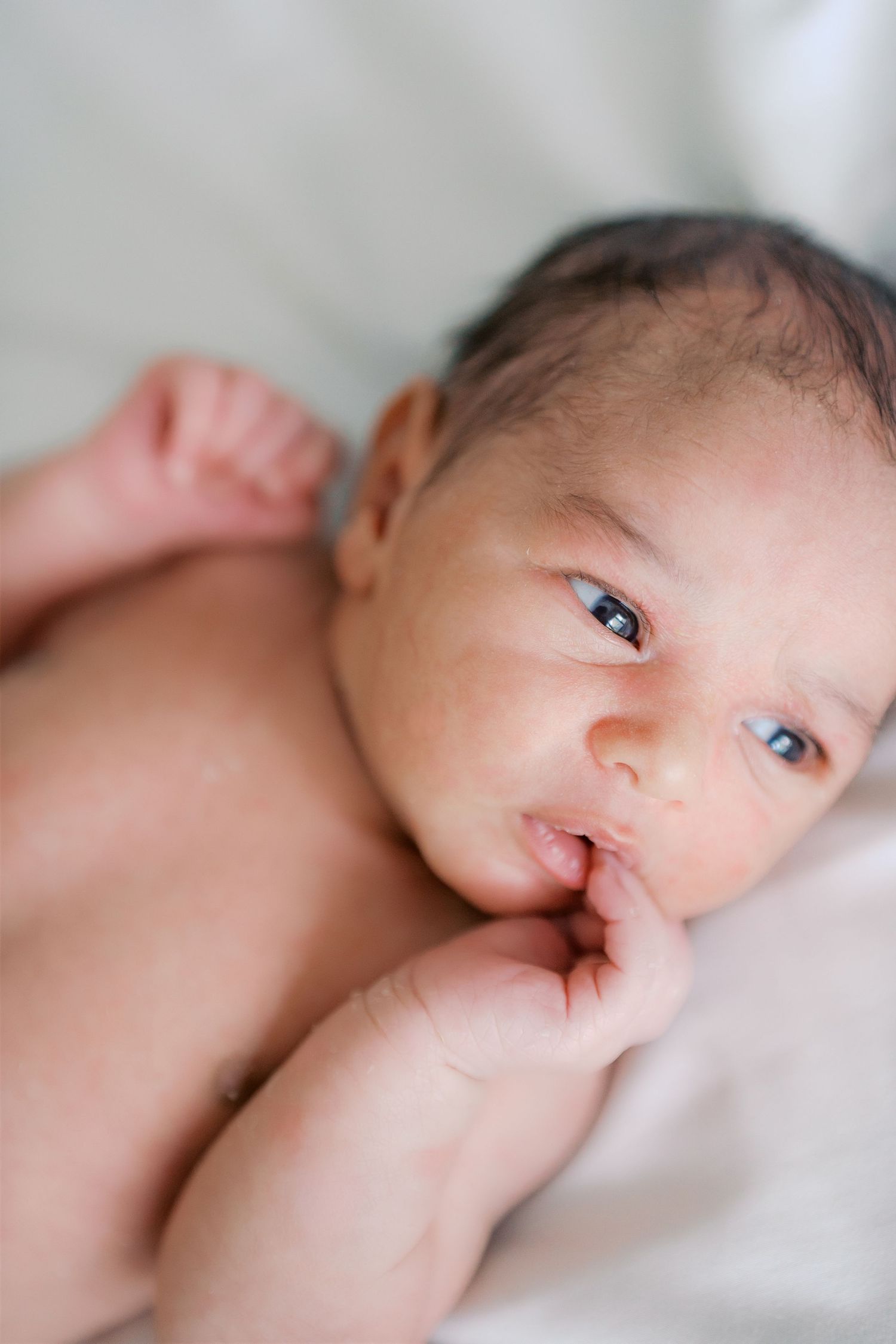 Newborn baby with dark hair and blue eyes lying on white blanket with finger in mouth.