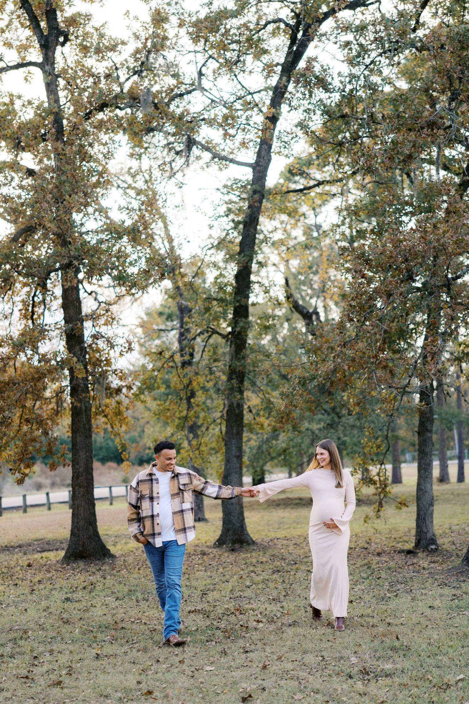 Couple holding hands in autumn park, pregnant woman in cream dress, man in plaid shirt and jeans, surrounded by fall trees.