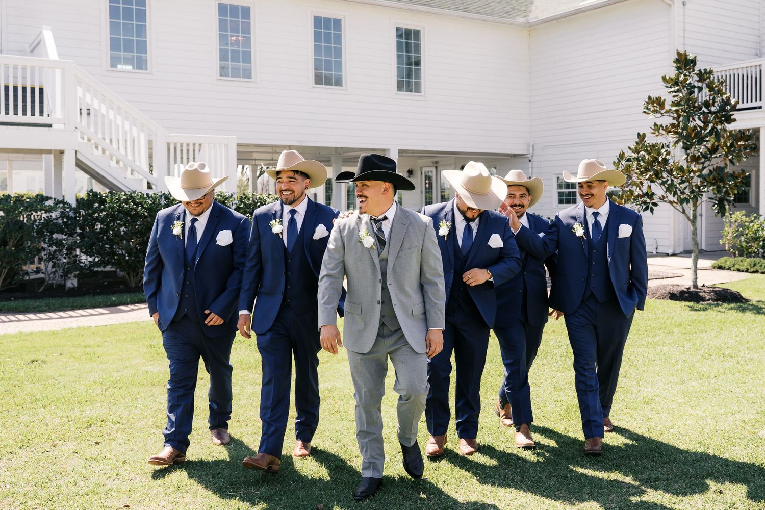 Wedding party in navy suits and gray attire pose in front of a white colonial building.