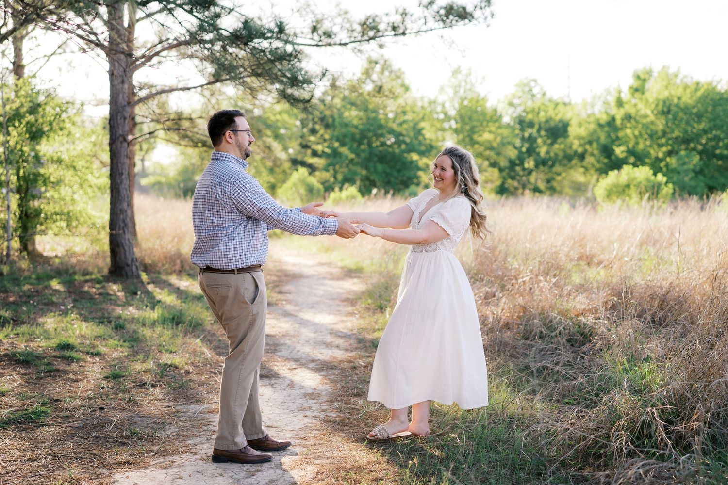Couple holding hands on a sunlit dirt path surrounded by golden grass and trees during an outdoor engagement session.