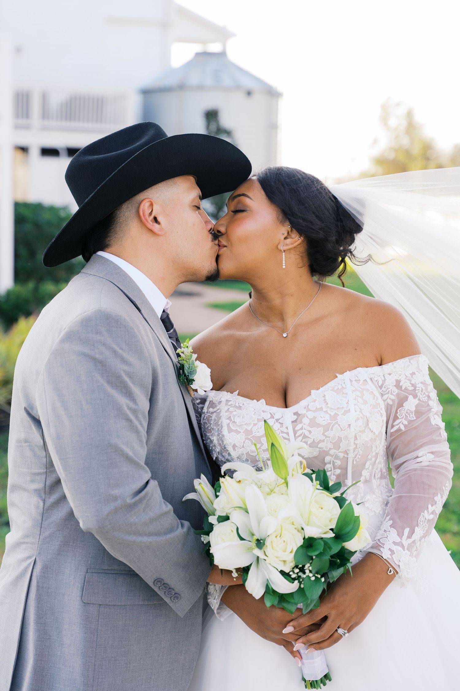 Wedding couple in gray suit and white lace dress share romantic kisses while holding white bouquet.