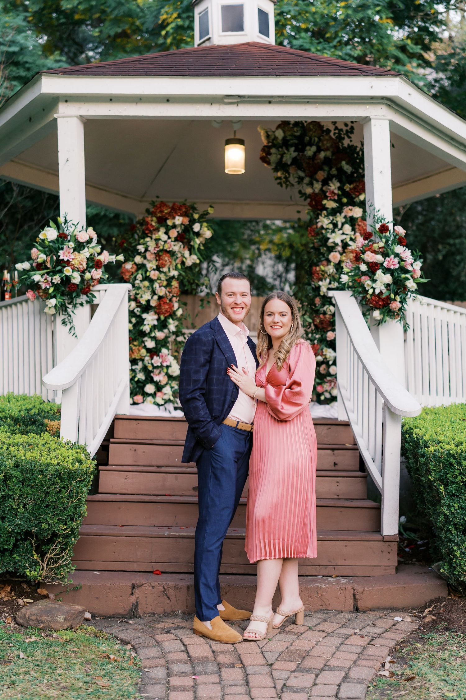 A couple poses together on wooden steps of a gazebo decorated with pink and white flowers.