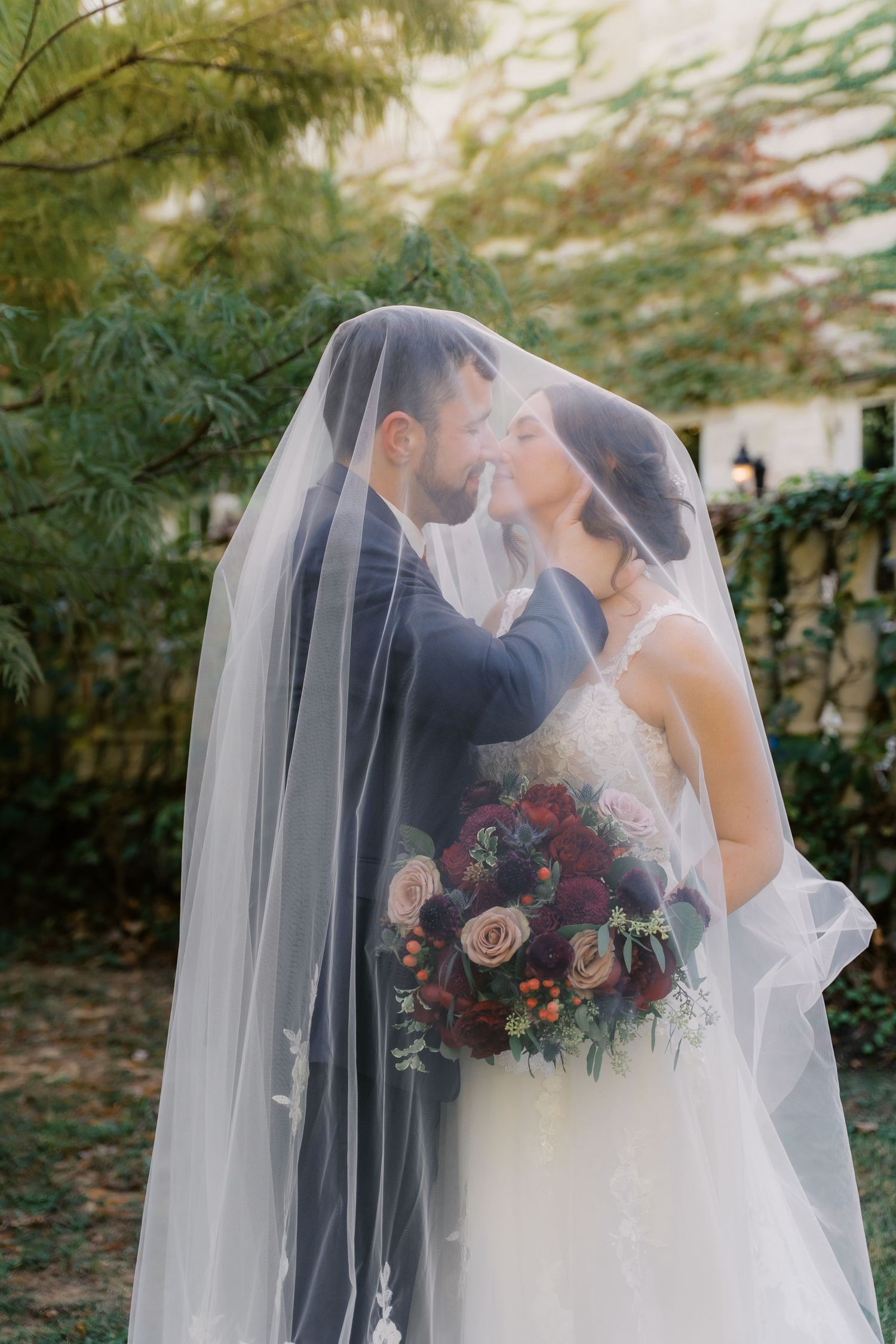 Romantic wedding couple embracing under a flowing veil in an outdoor garden setting.