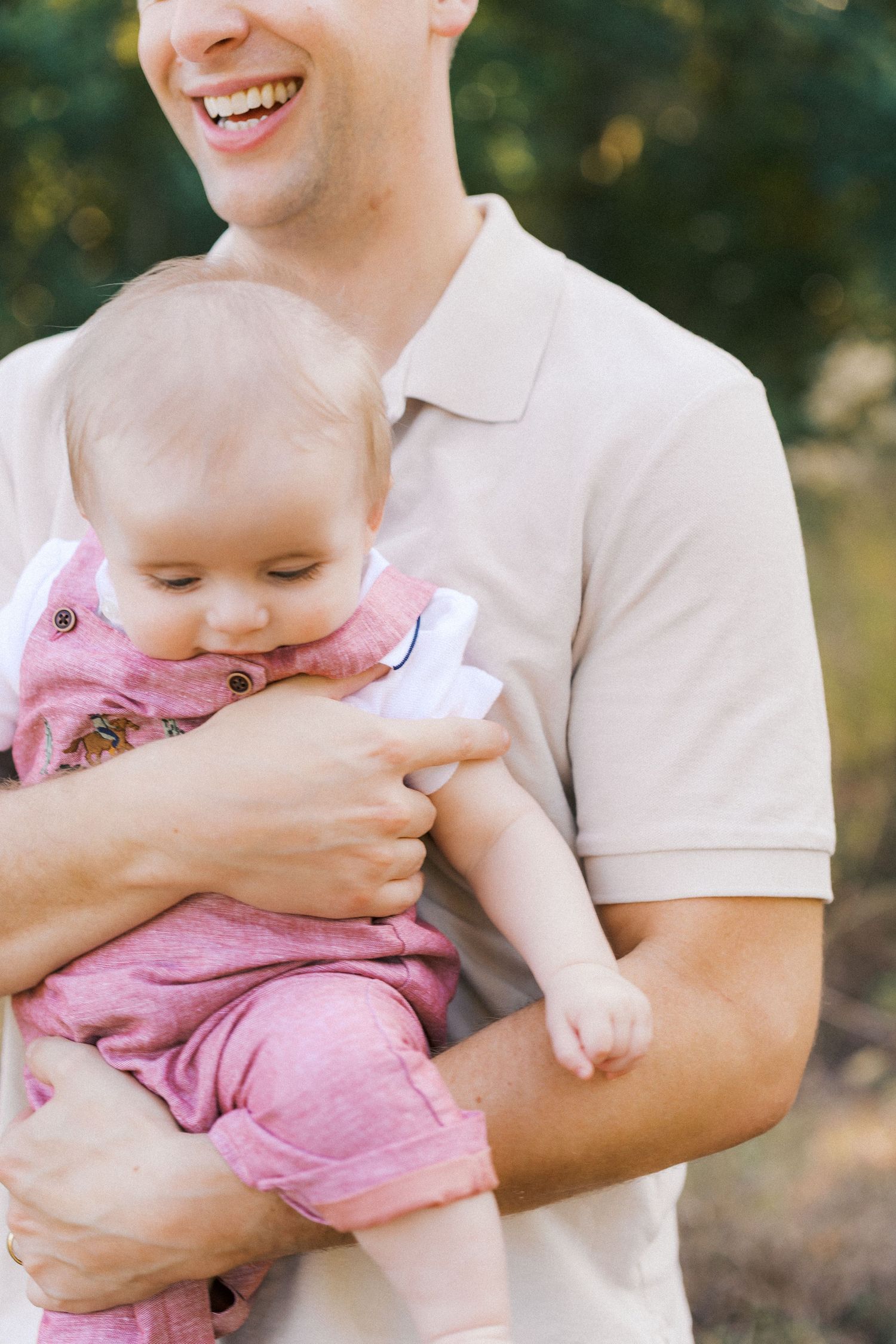 A baby wearing a pink romper is held lovingly in someone's arms in an outdoor setting.