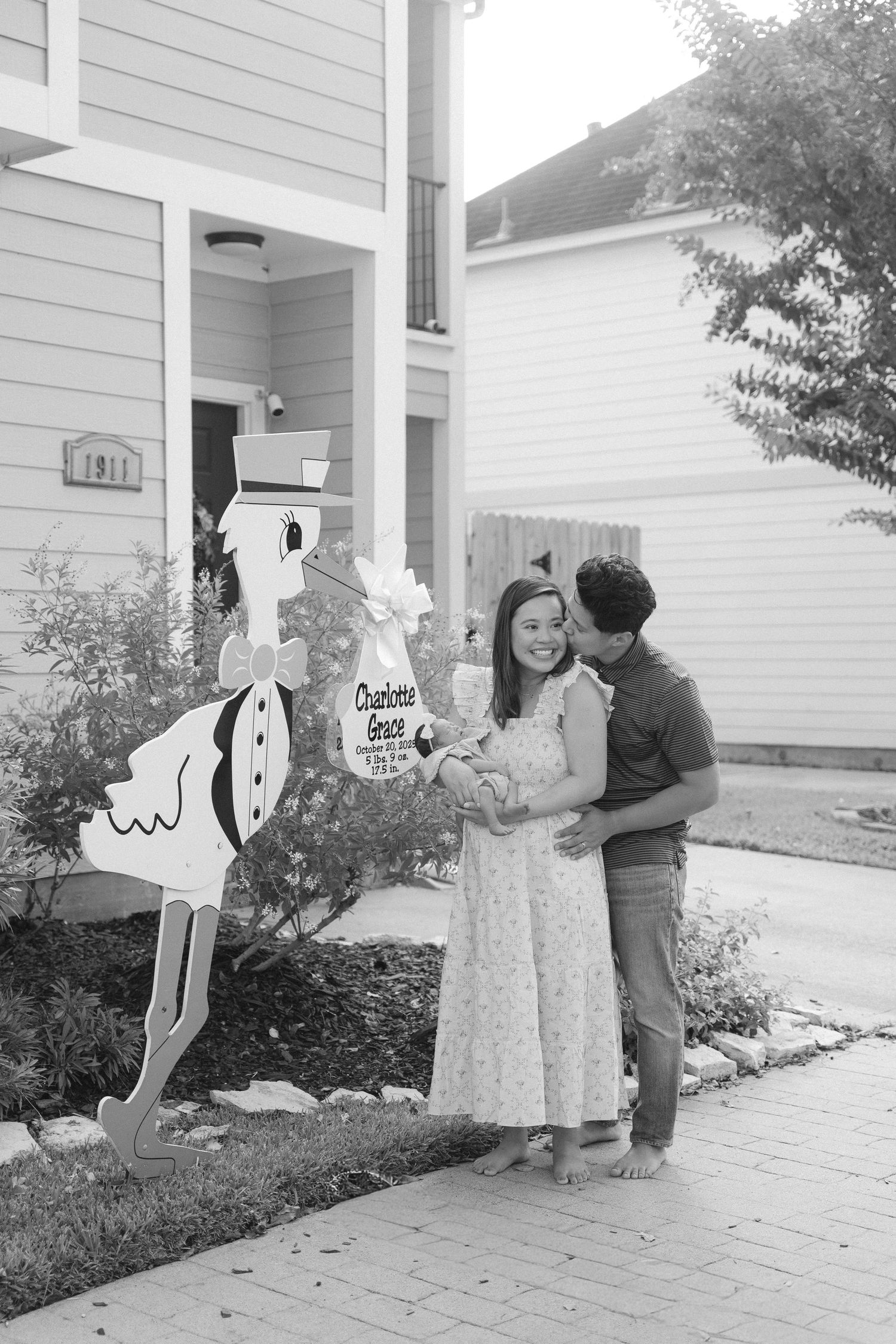 Black and white photo of family gathering by stork yard sign outside their home.