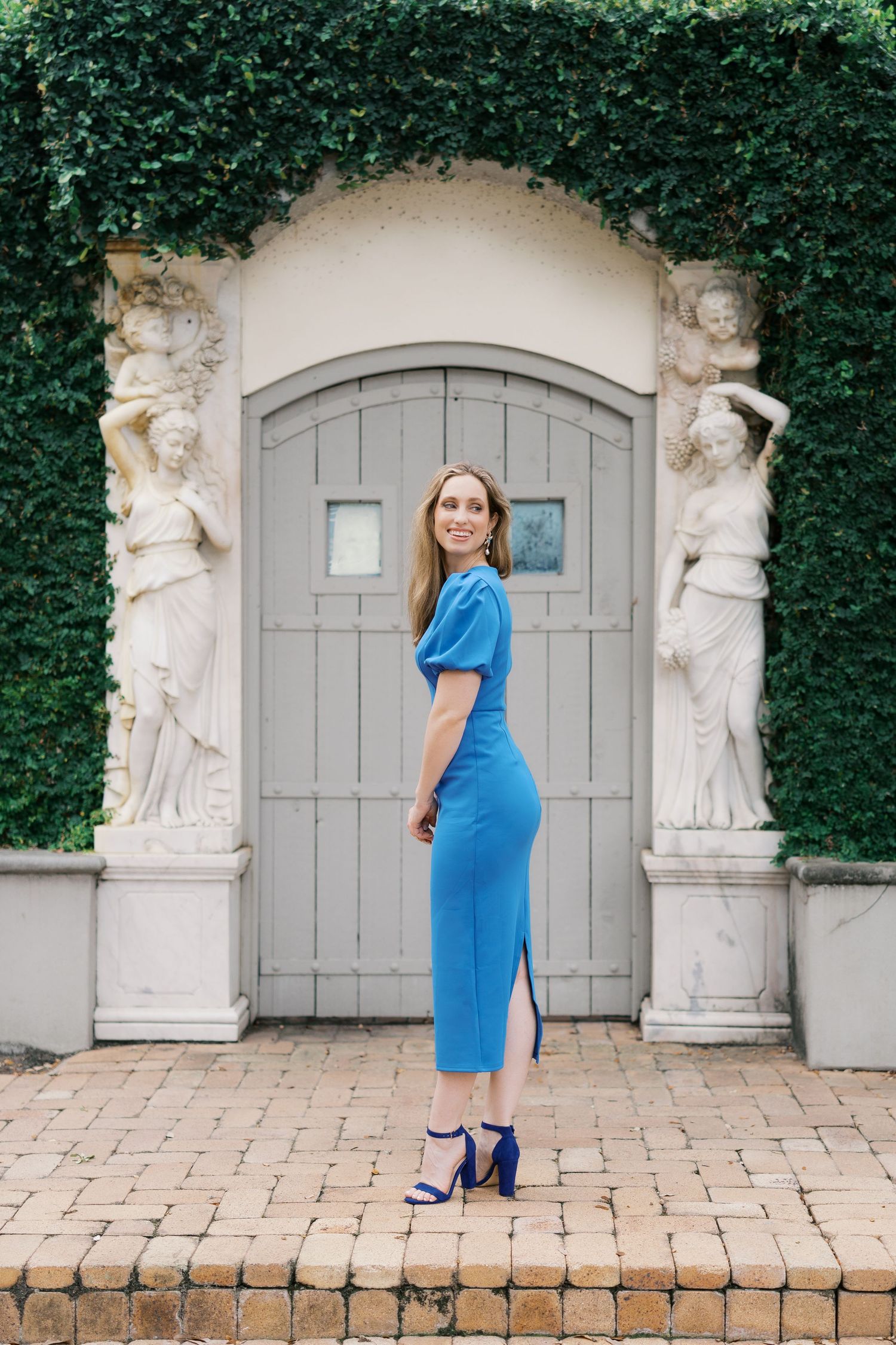 Person in bright blue dress poses in front of ornate garden archway with classical statues.