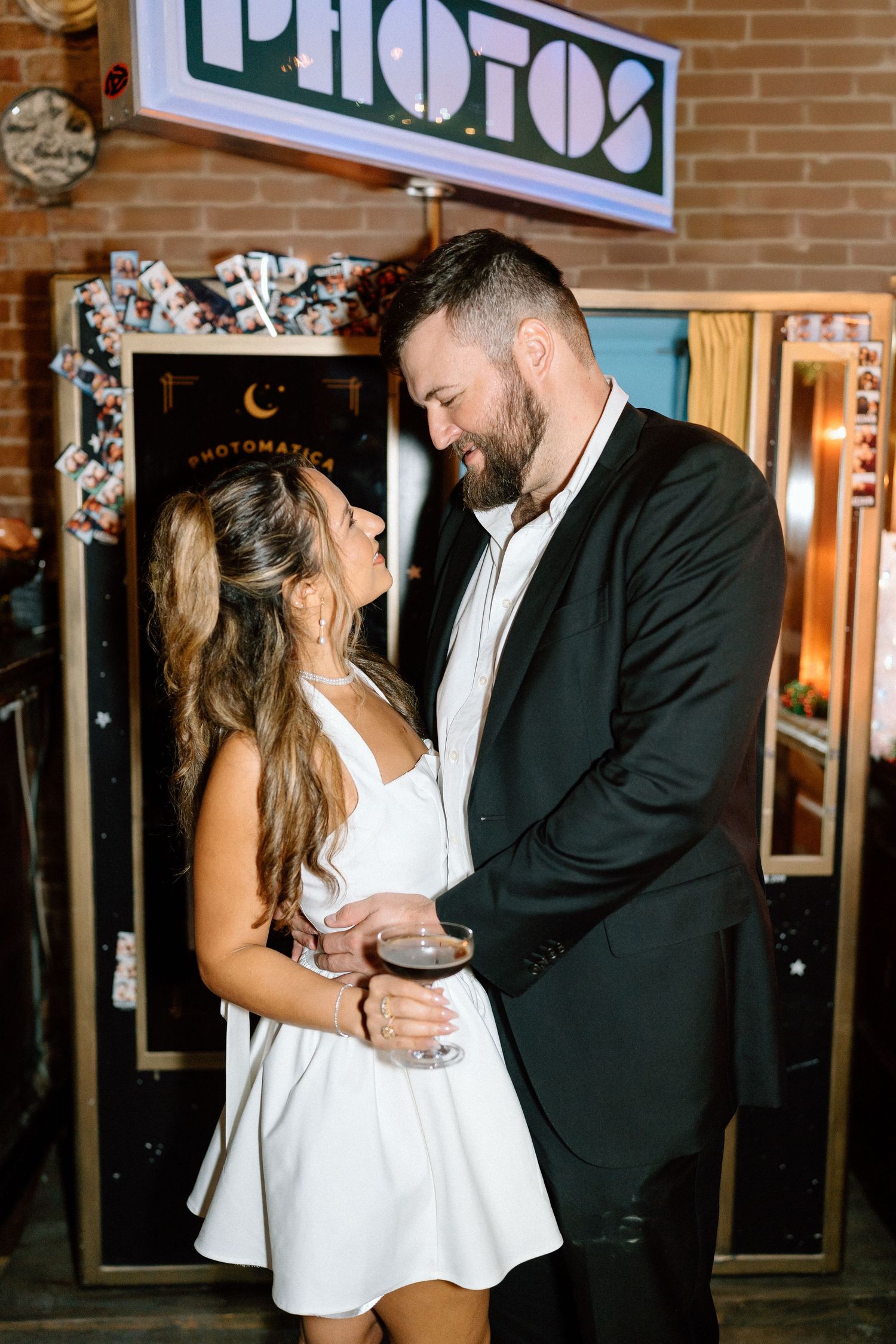 Romantic couple shares intimate moment at bar with exposed brick wall and neon lighting in background.