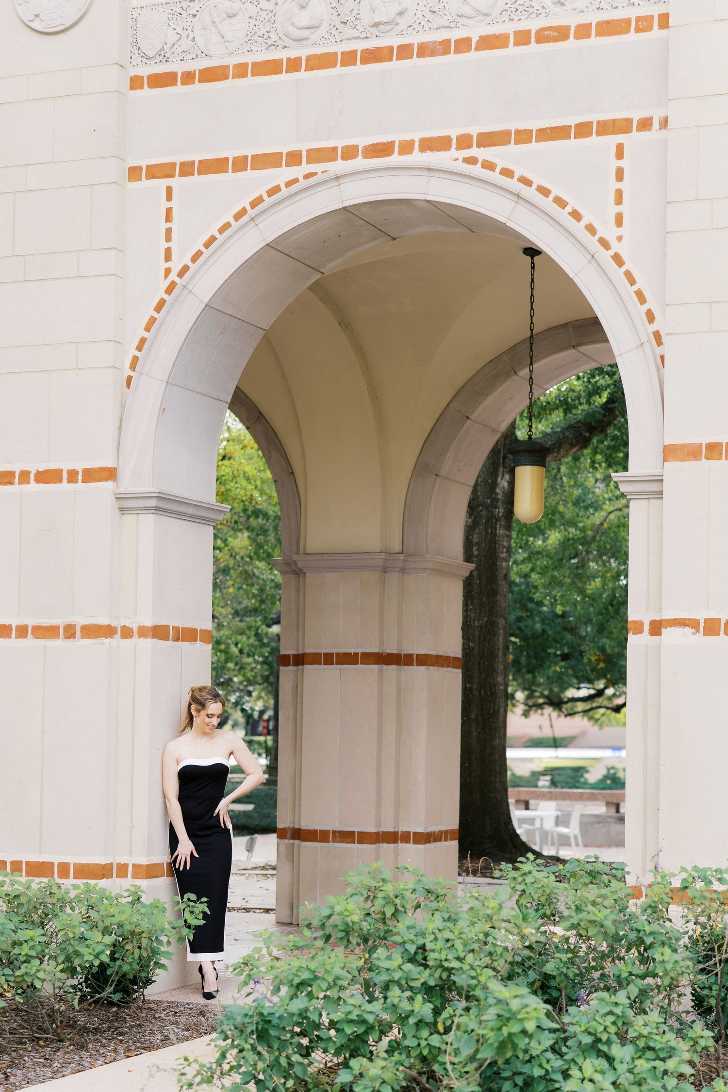 A woman in a black strapless jumpsuit poses under an ornate brick archway surrounded by lush greenery.