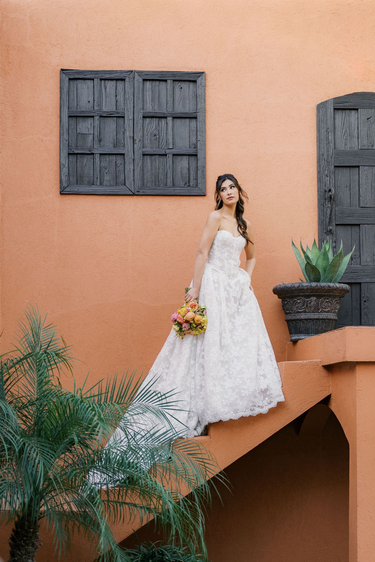 Bride in white lace ball gown holding colorful bouquet stands on terrace of orange Spanish-style building.