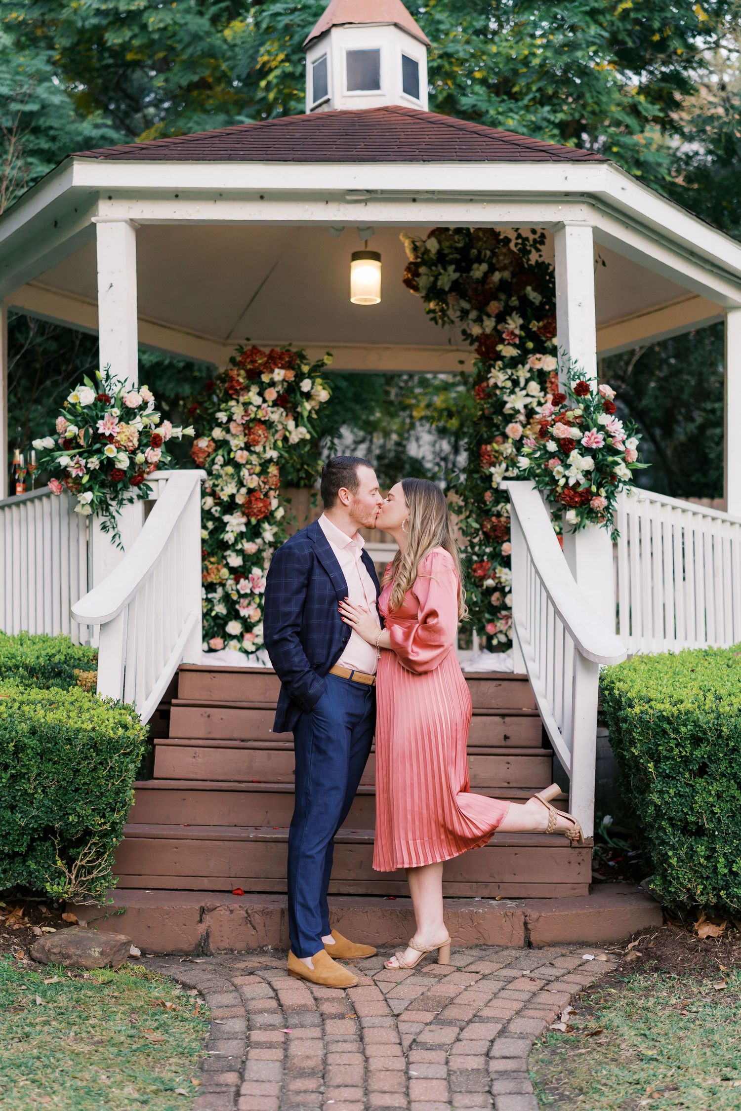 A couple embraces on gazebo steps surrounded by pink and white floral arrangements.