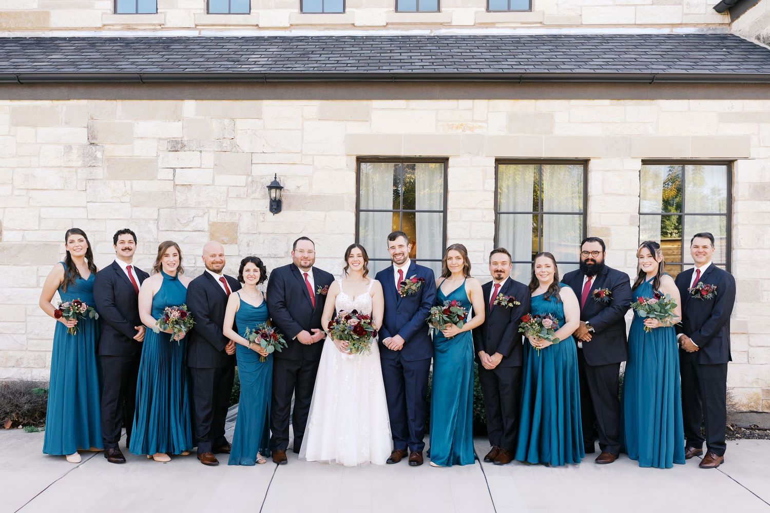 Wedding party in teal dresses and navy suits posed along stone building exterior.
