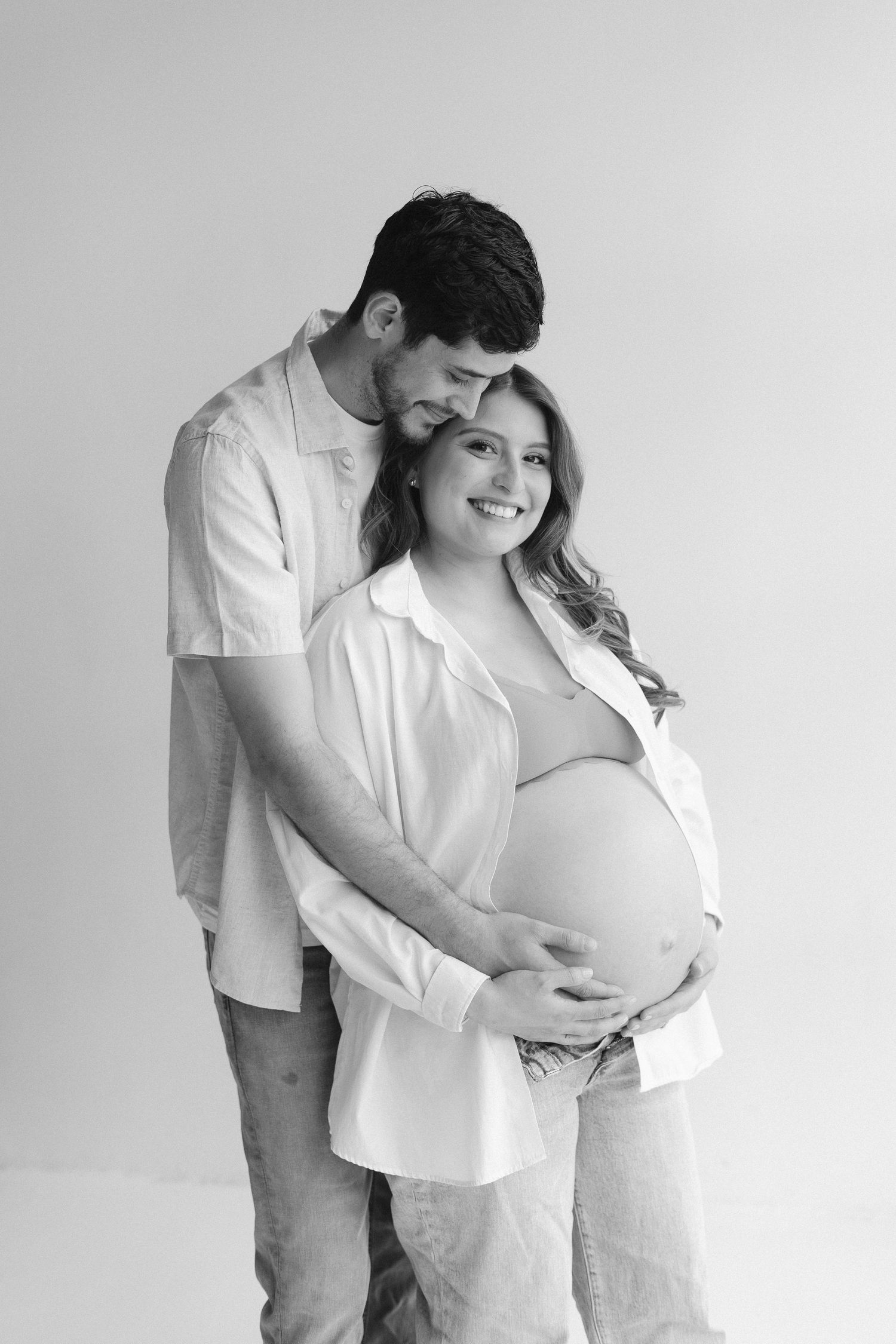 Maternity photo series showing couple in white shirts embracing against light background.