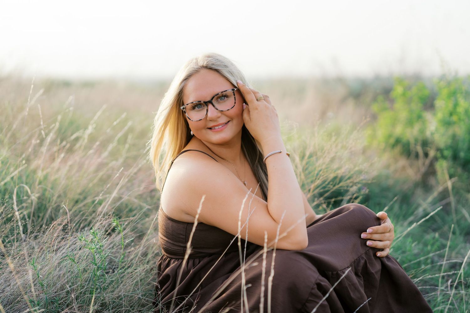 Smiling blonde woman with glasses sitting in tall grass field wearing brown dress in golden hour light.