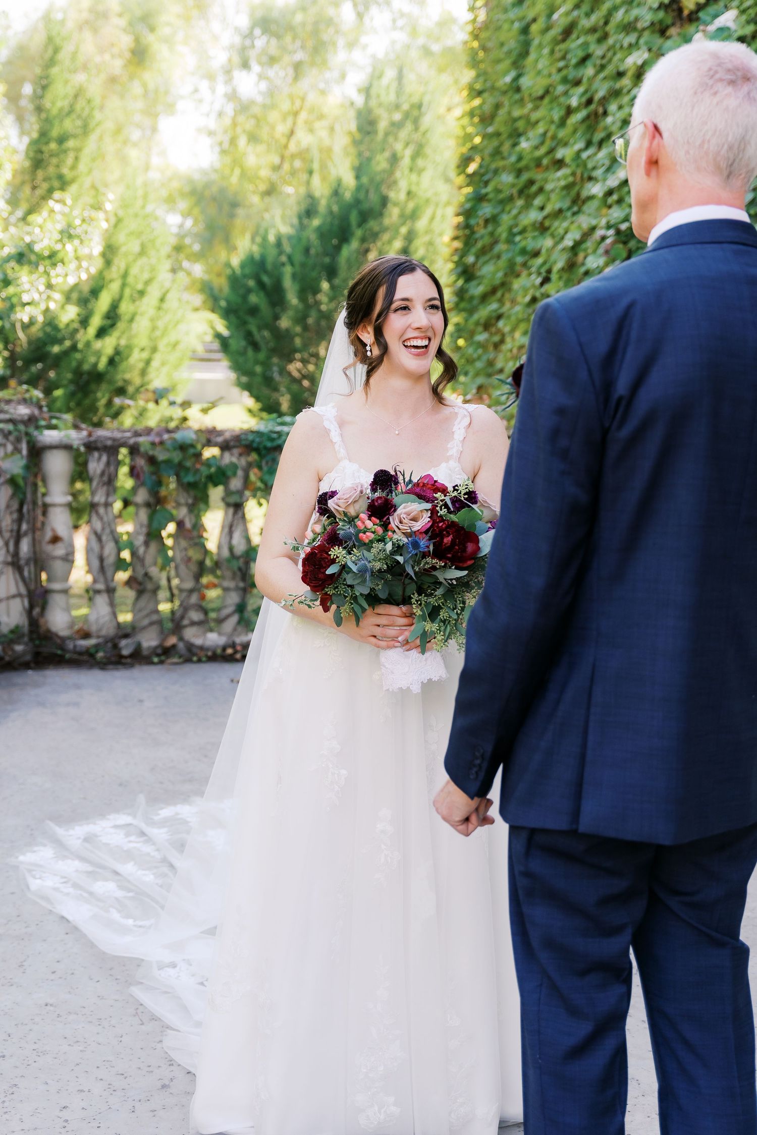 Bride in white gown holding burgundy bouquet shares a tender moment with wedding guest in navy suit outdoors.
