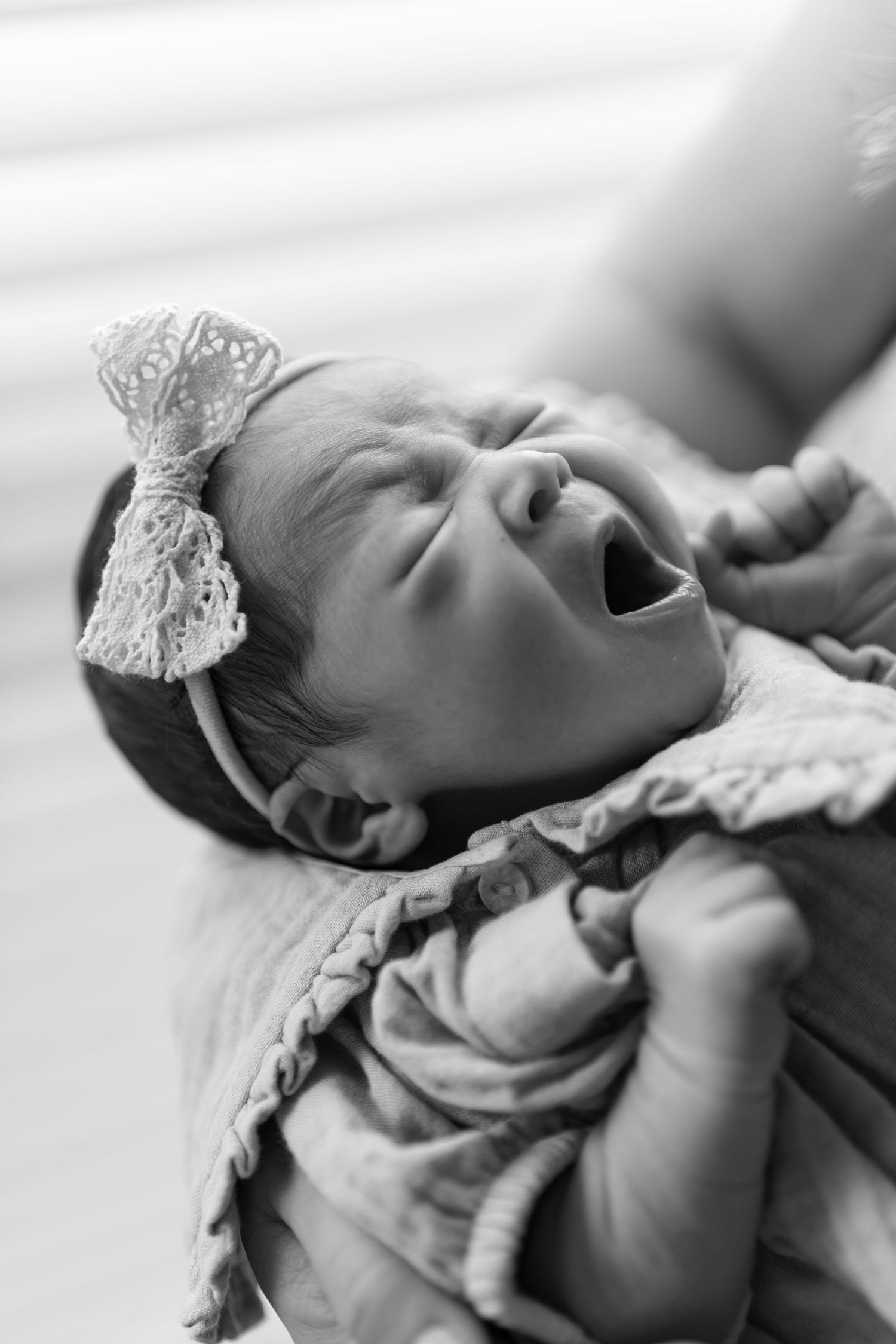 Black and white photo of a yawning newborn baby wearing a lace bow headband.