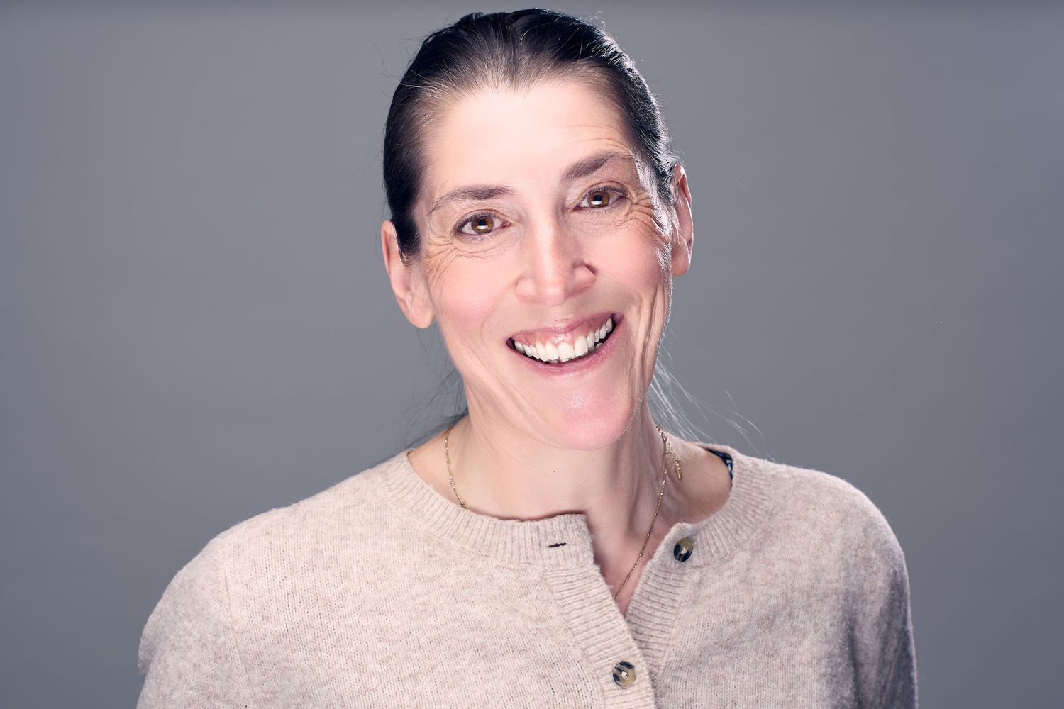 A smiling person wearing glasses and a beige sweater poses against a gray background.