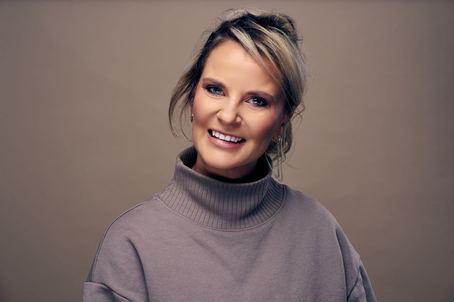 A person wearing a gray turtleneck sweater smiles warmly against a neutral background.