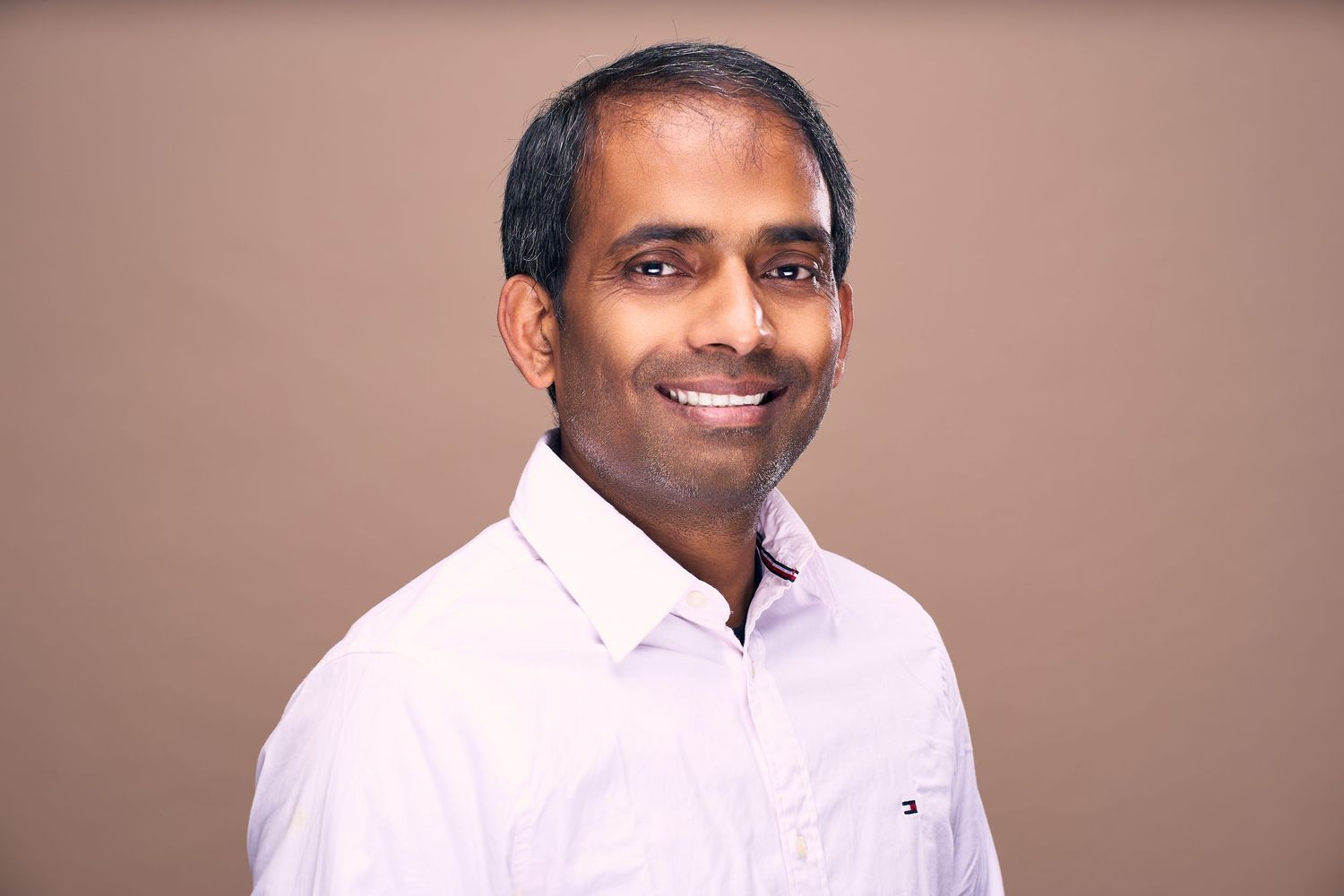 Professional headshot of someone in a white button-down shirt smiling warmly against a neutral brown background.