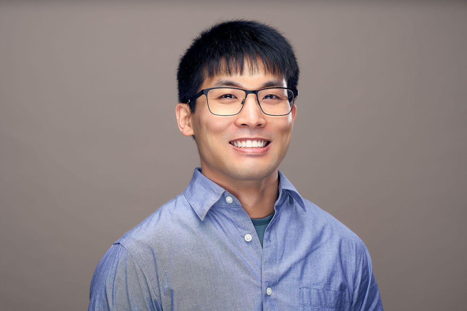 Professional headshot of person wearing glasses and blue button-down shirt against neutral background.
