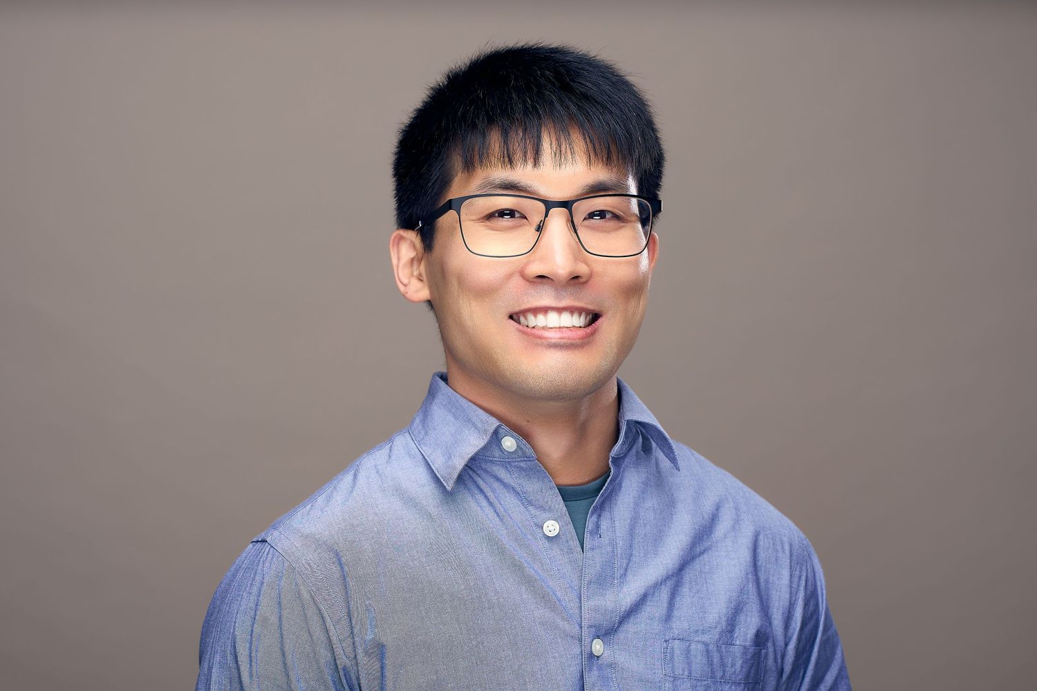 Professional headshot of someone wearing glasses and a light blue button-down shirt against a neutral background.