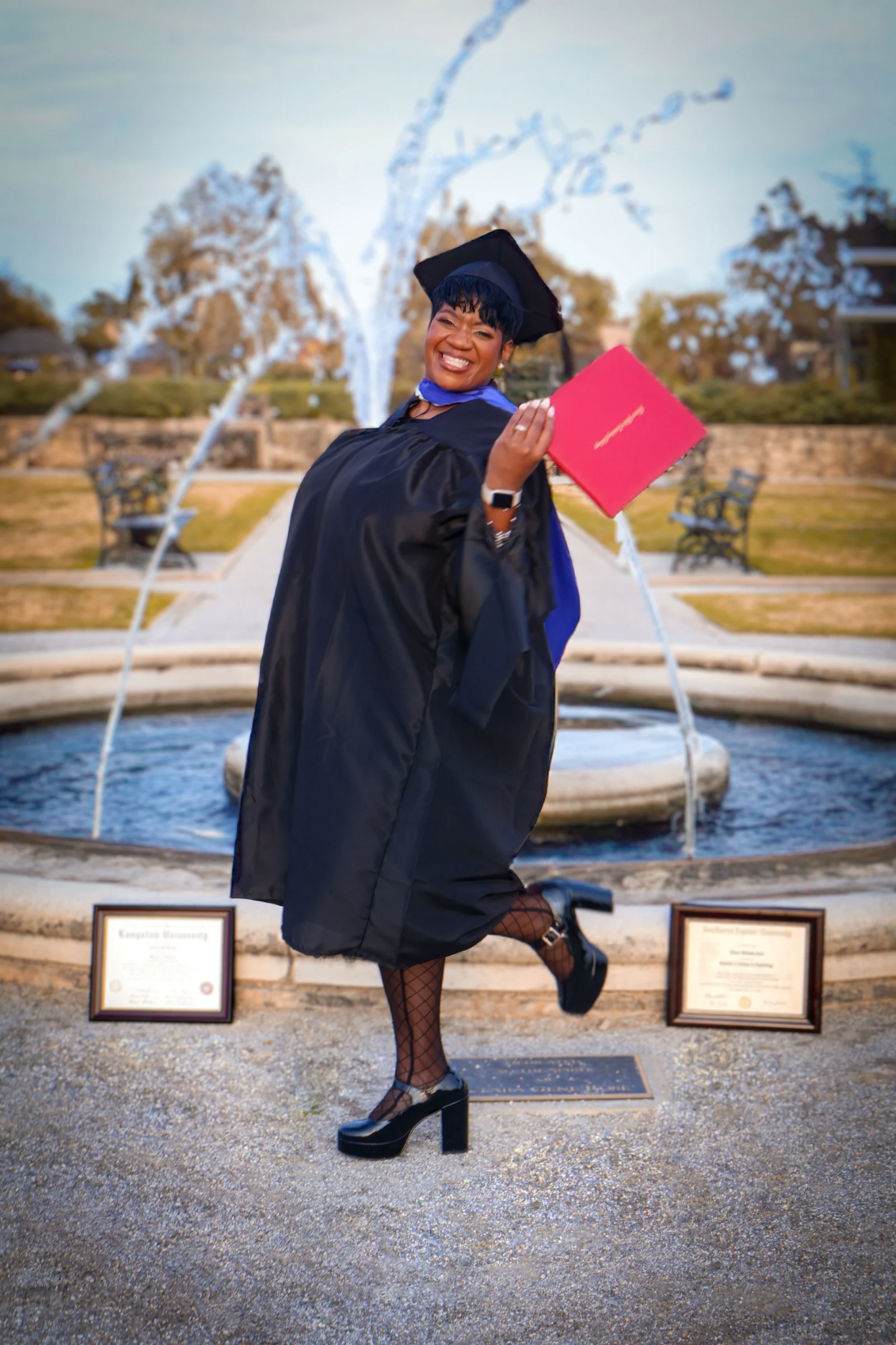Joyful Black woman in graduation cap and gown holds diploma by fountain with award plaques.
