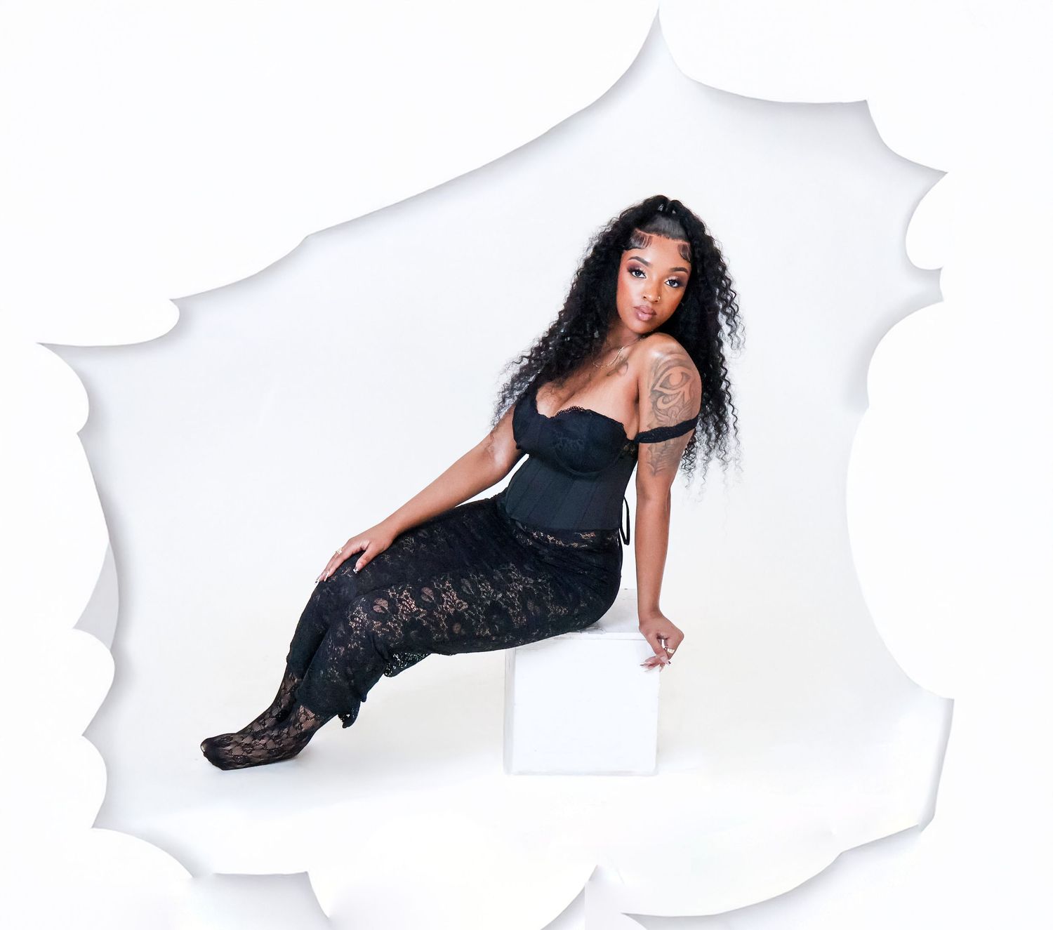 Woman in black lace outfit sitting on white cube cutout.