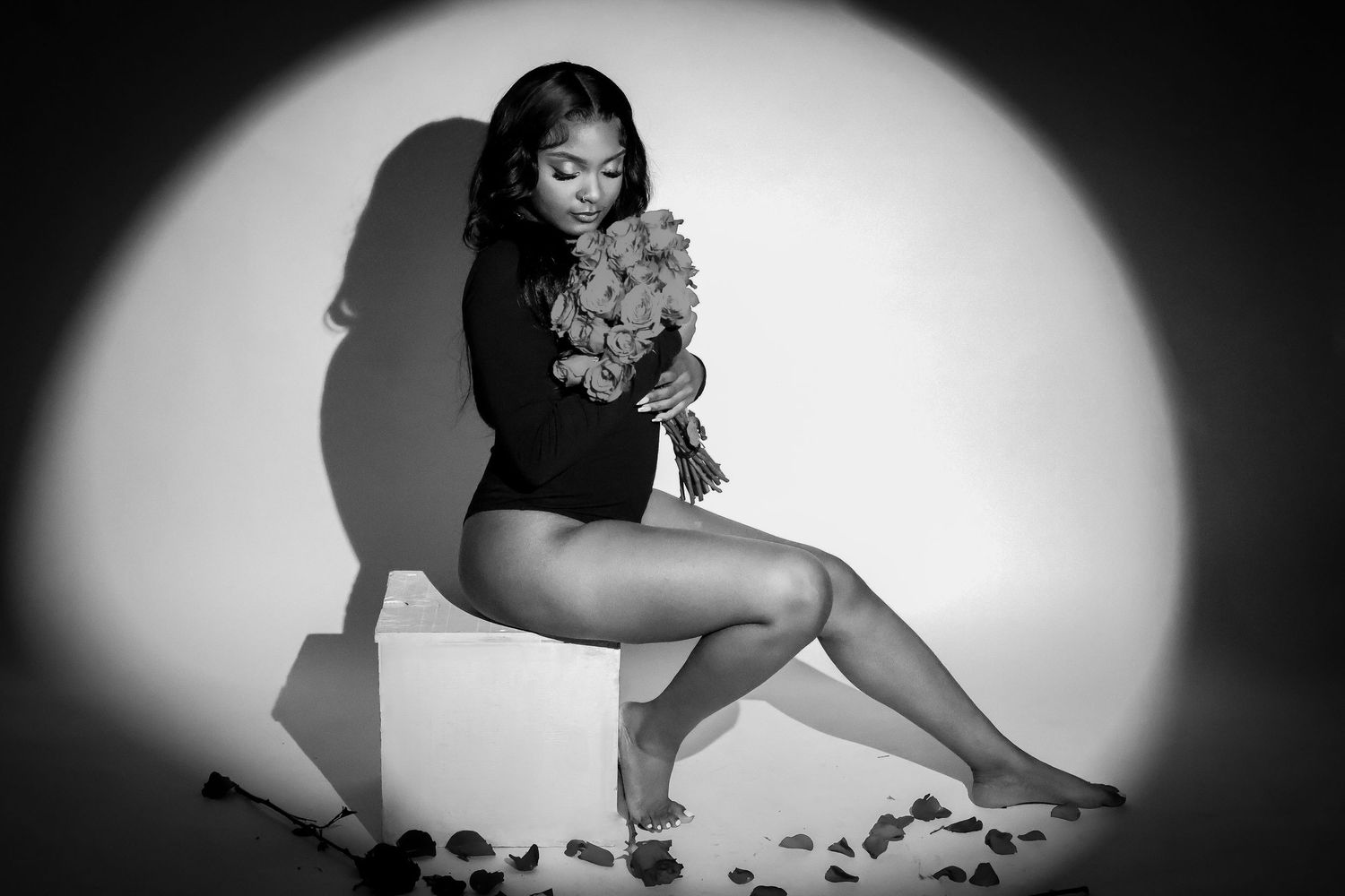 Black and white studio photo of woman in black bodysuit holding bouquet of roses on cube.