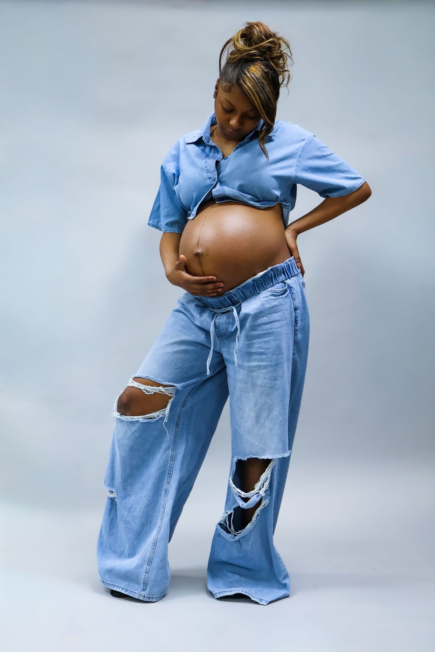 Pregnant woman in blue crop top and ripped wide-leg jeans cradling her baby bump against gray background.