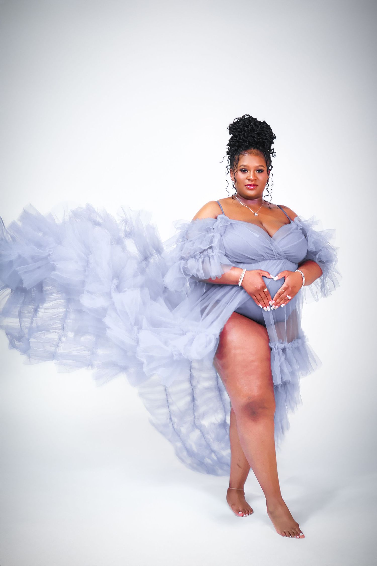 Pregnant woman in flowing gray tulle gown poses gracefully, forming heart shape with hands on baby bump.