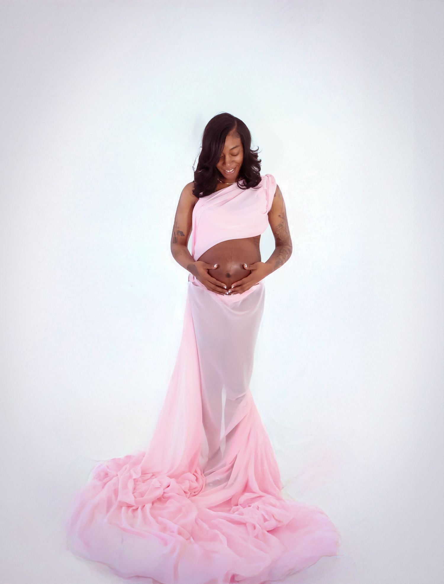 Pregnant woman in elegant pink chiffon maternity gown cradling baby bump during professional studio photoshoot.