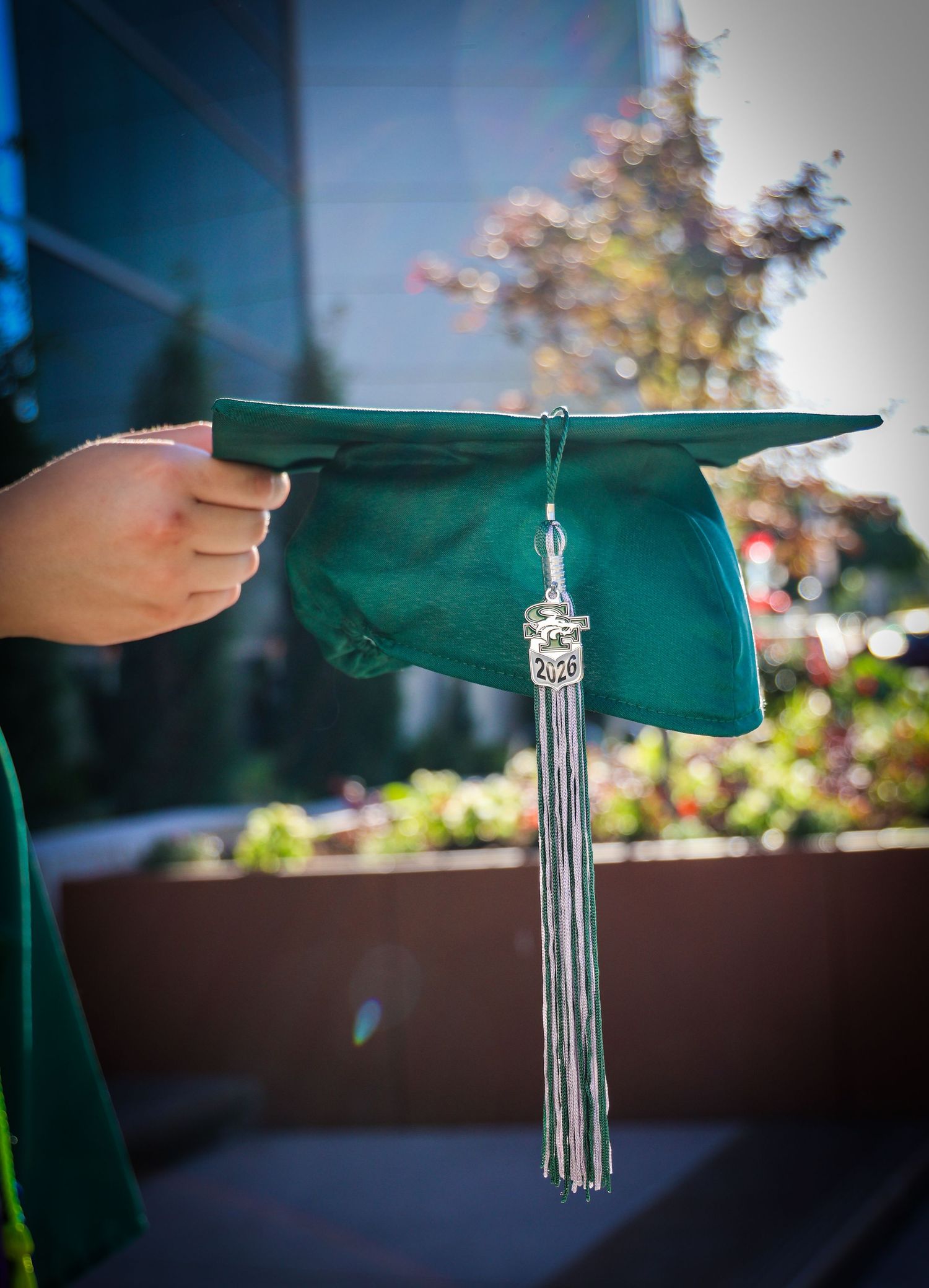 A hand holding a green graduation cap with a silver tassel against a blurred outdoor background.