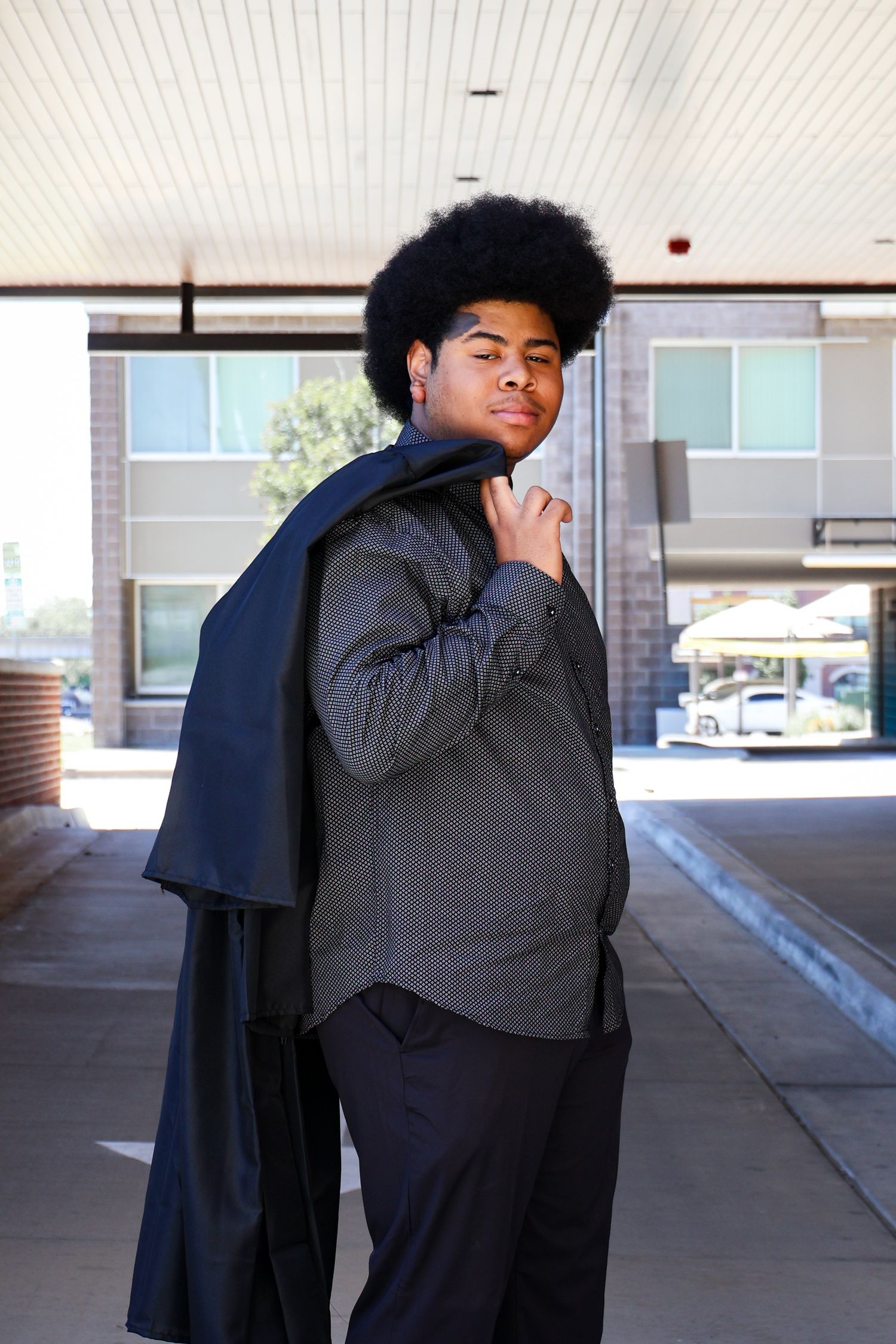 Young Black man with afro hair drapes suit jacket over shoulder outdoors near brick building in stylish casual pose.