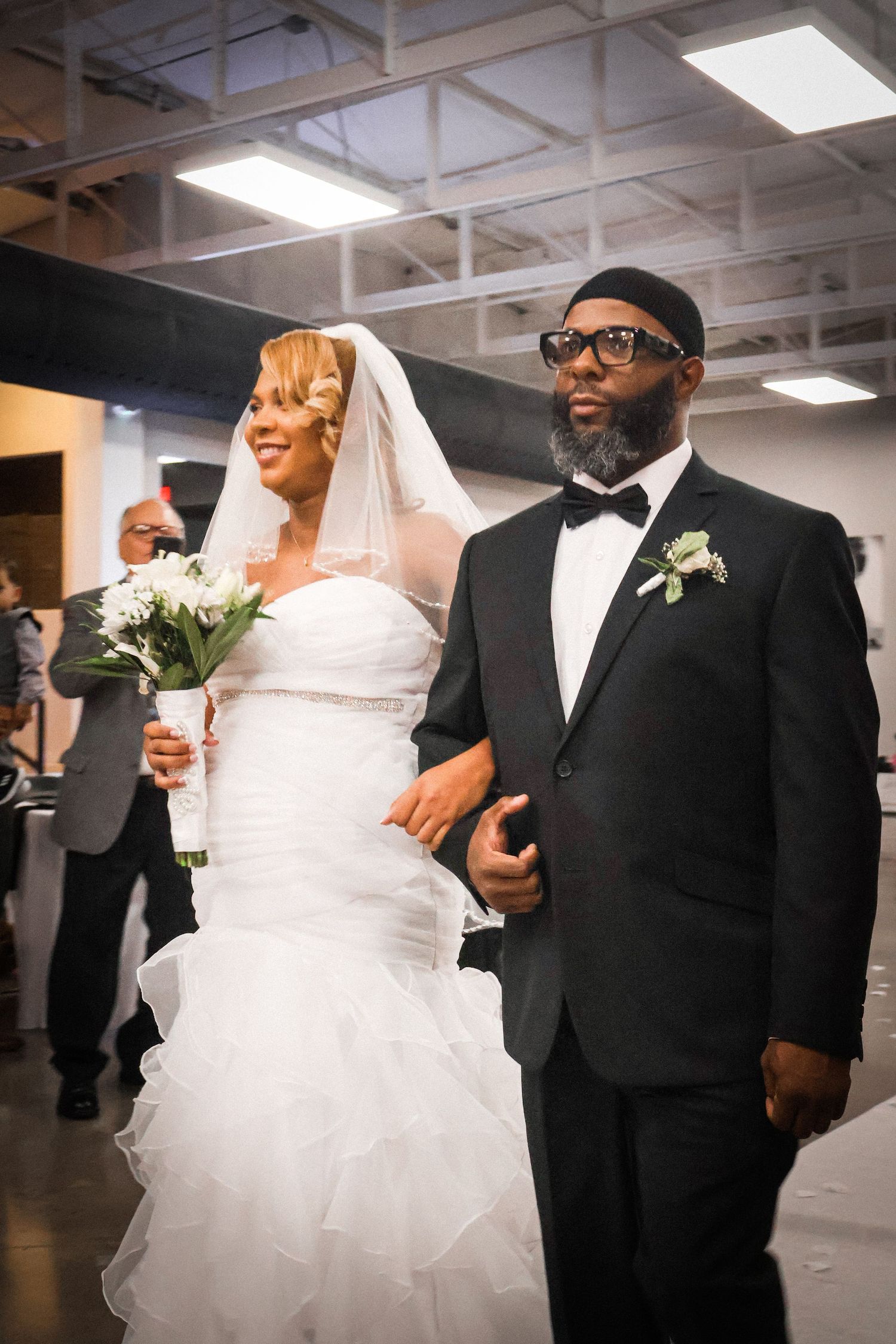 Bride in white strapless gown with veil and bouquet walks with groom in black tuxedo and bow tie at wedding ceremony.