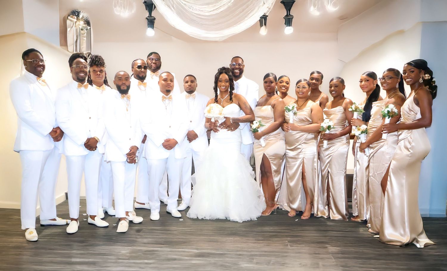 Wedding party poses together, groomsmen in white suits and bridesmaids in gold gowns flanking bride and groom.