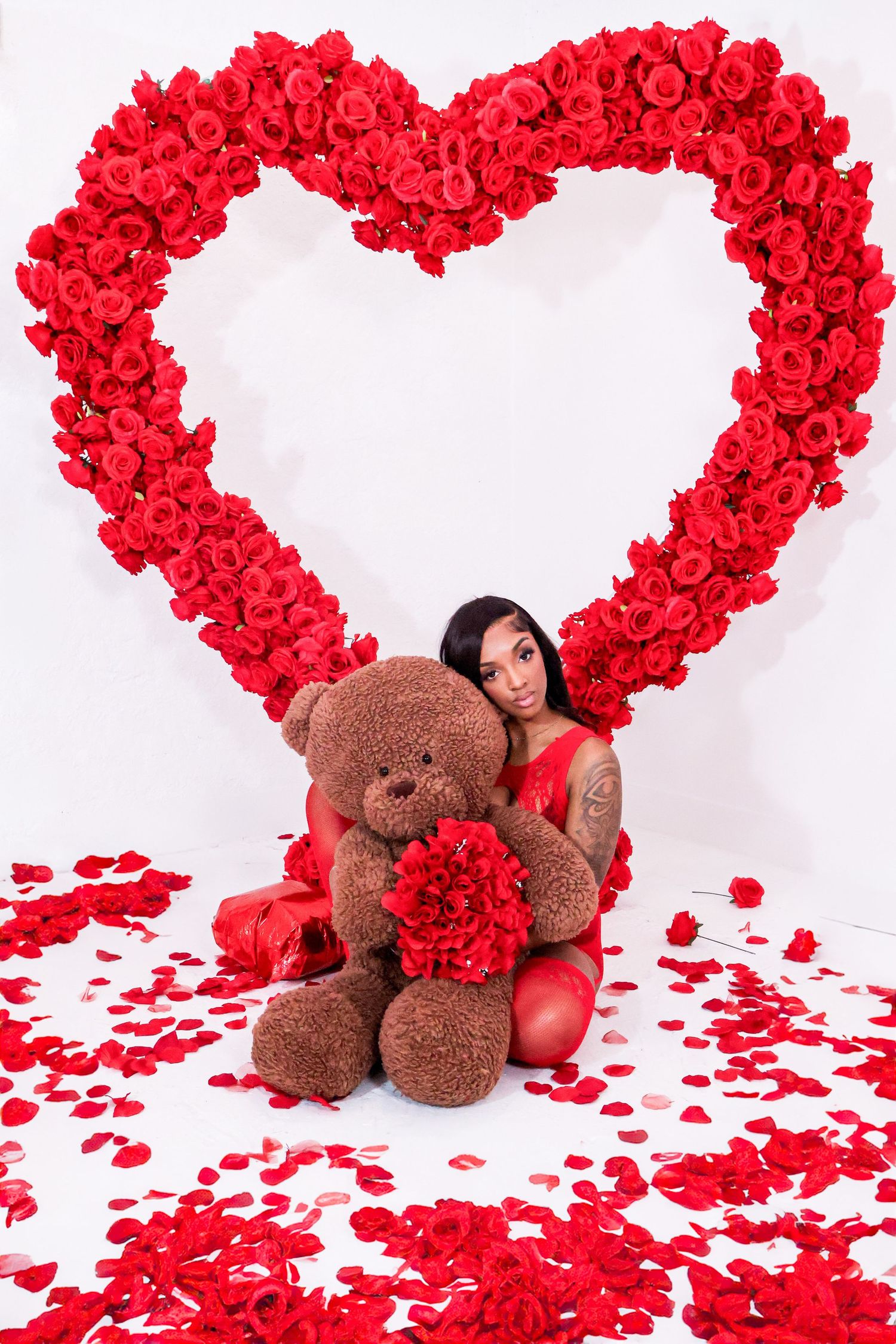 Woman in red dress hugging teddy bear in front of red rose heart backdrop.