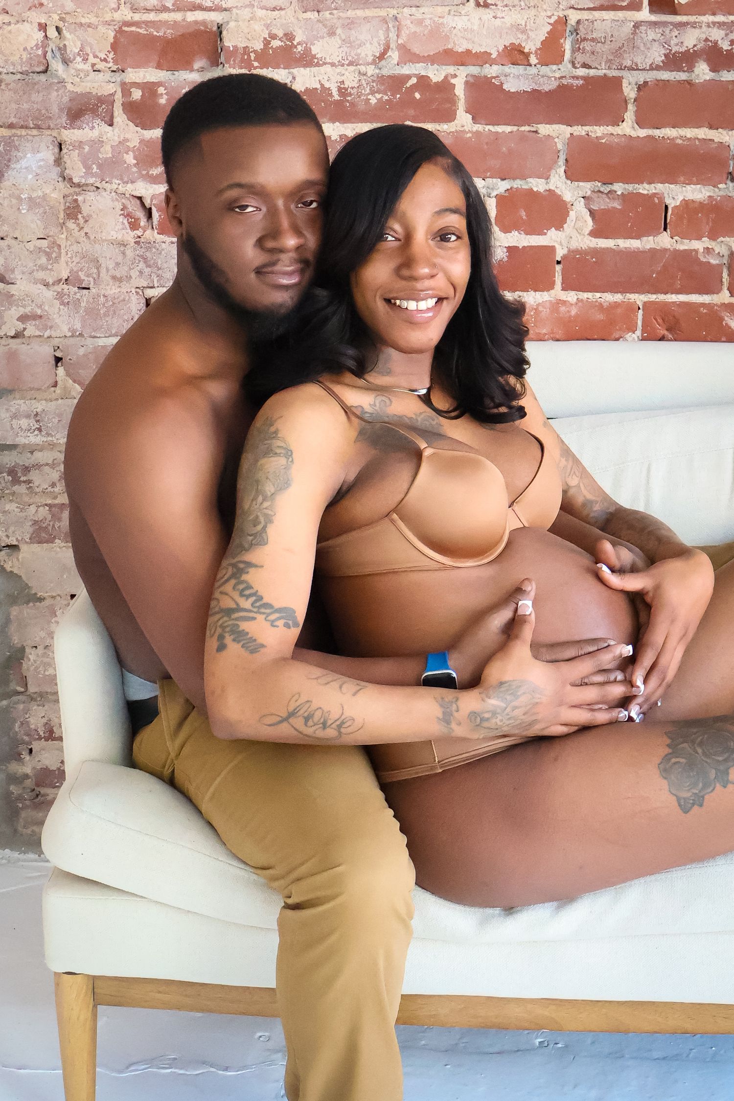 A happy couple poses for a maternity photo shoot, with the man embracing the pregnant woman in front of a brick wall.